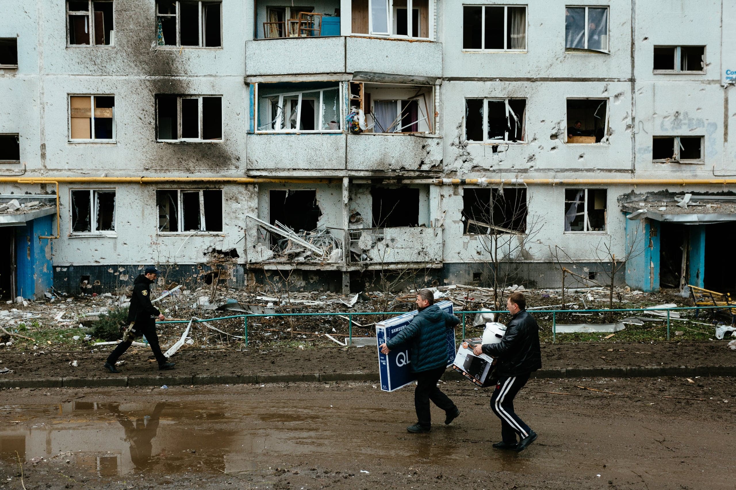 Разрушенный обстрелами фасад жилого дома. Двое мужчин несут по грязной дороге коробки с бытовой техникой, на одной из которых написано "QLED". Слева проходит человек в темной форме.