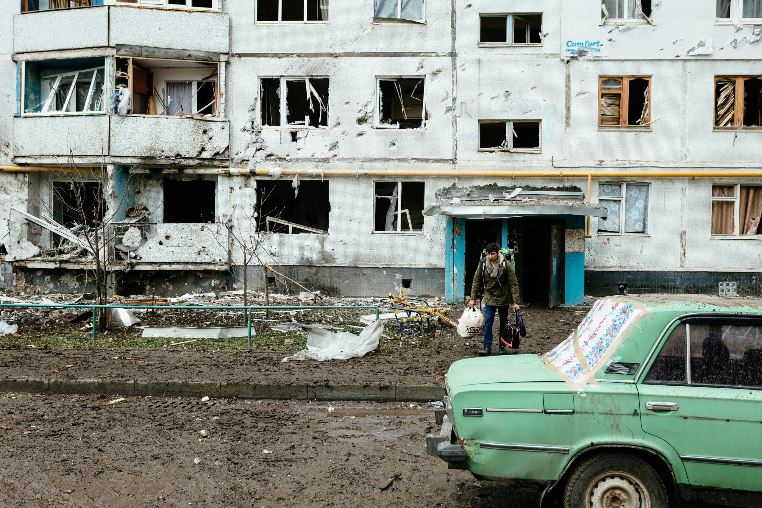 Разрушенный обстрелами фасад многоквартирного дома с выбитыми окнами и осколочными следами. Мужчина с рюкзаком и сумками выходит из подъезда. На переднем плане старый зеленый автомобиль на грязной дороге. На фасаде видна надпись "Comfort".