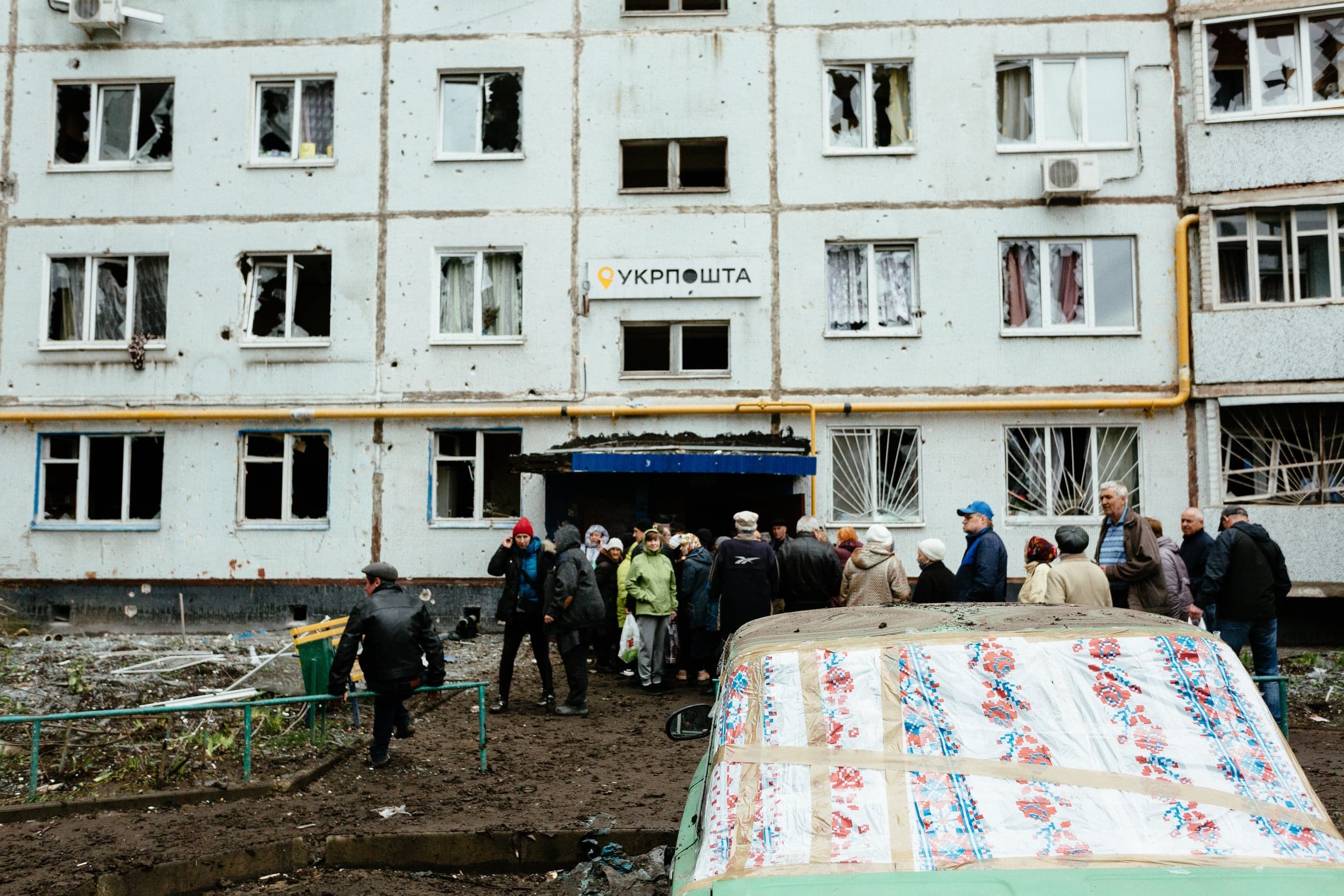 Поврежденный обстрелом жилой дом с выбитыми окнами и вывеской "Укрпошта". Толпа людей собралась у входа; на переднем плане — укрытый автомобиль.