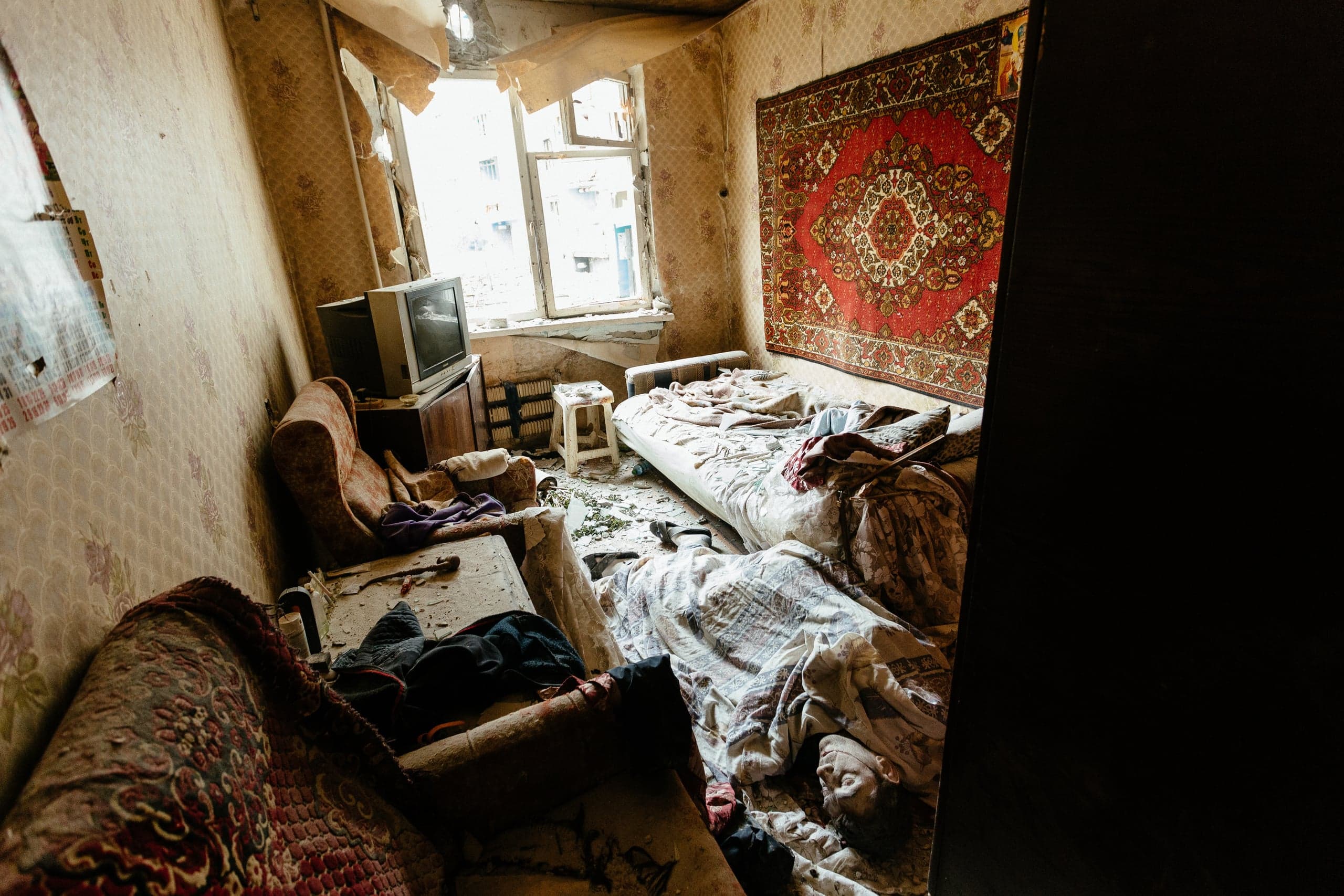 Разрушенная комната с порванными обоями, красным ковром на стене, разбитым окном, старым телевизором, разбросанными вещами и телом человека, лежащим на полу.