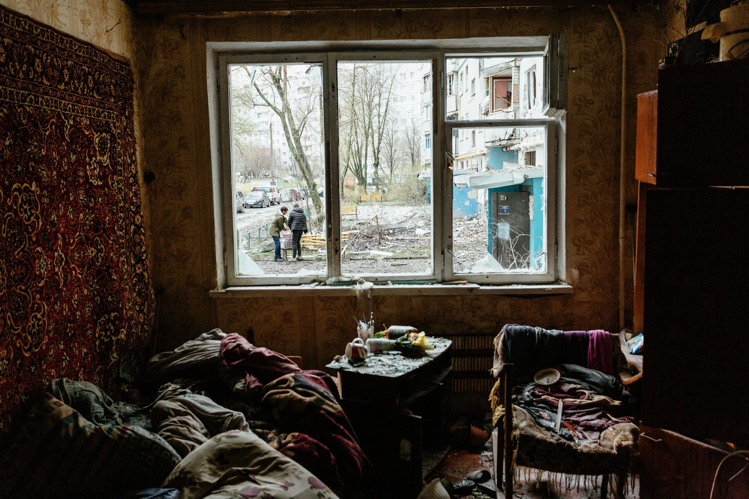 Поврежденная комната с ковром на стене; вид через разбитое окно на разрушенный двор, где стоят двое людей.