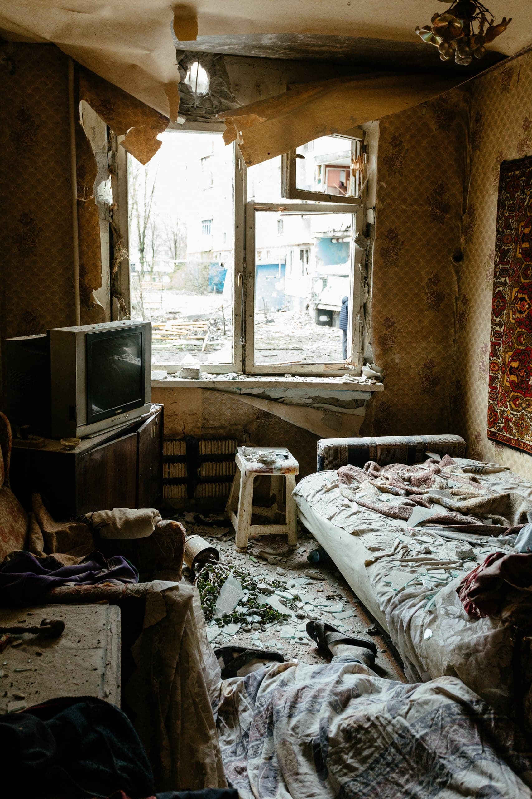 Интерьер разрушенной комнаты: разбитое окно, оторванные обои, осколки на полу и кровати, старый телевизор.