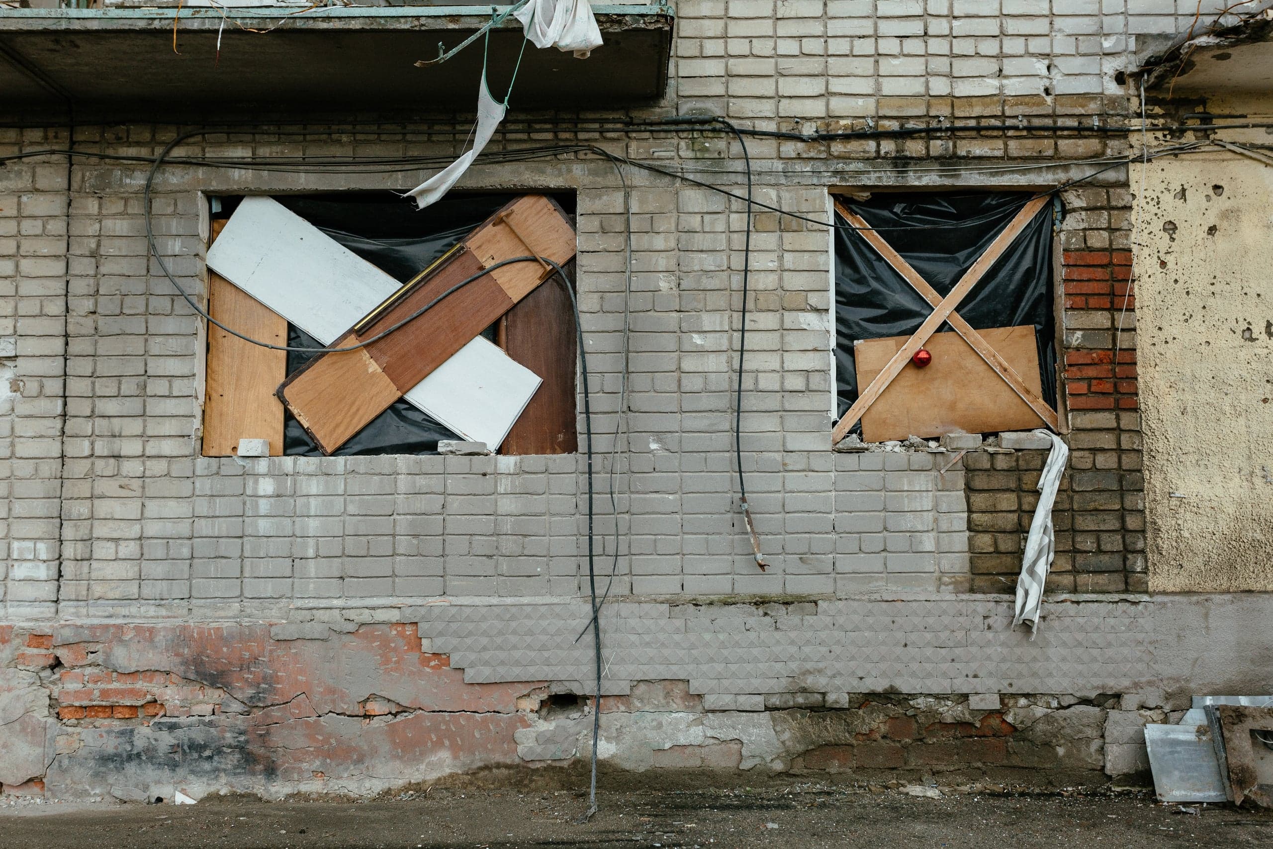 Поврежденный кирпичный фасад здания с двумя окнами, заколоченными обломками дерева, фанерой и черной пленкой.
