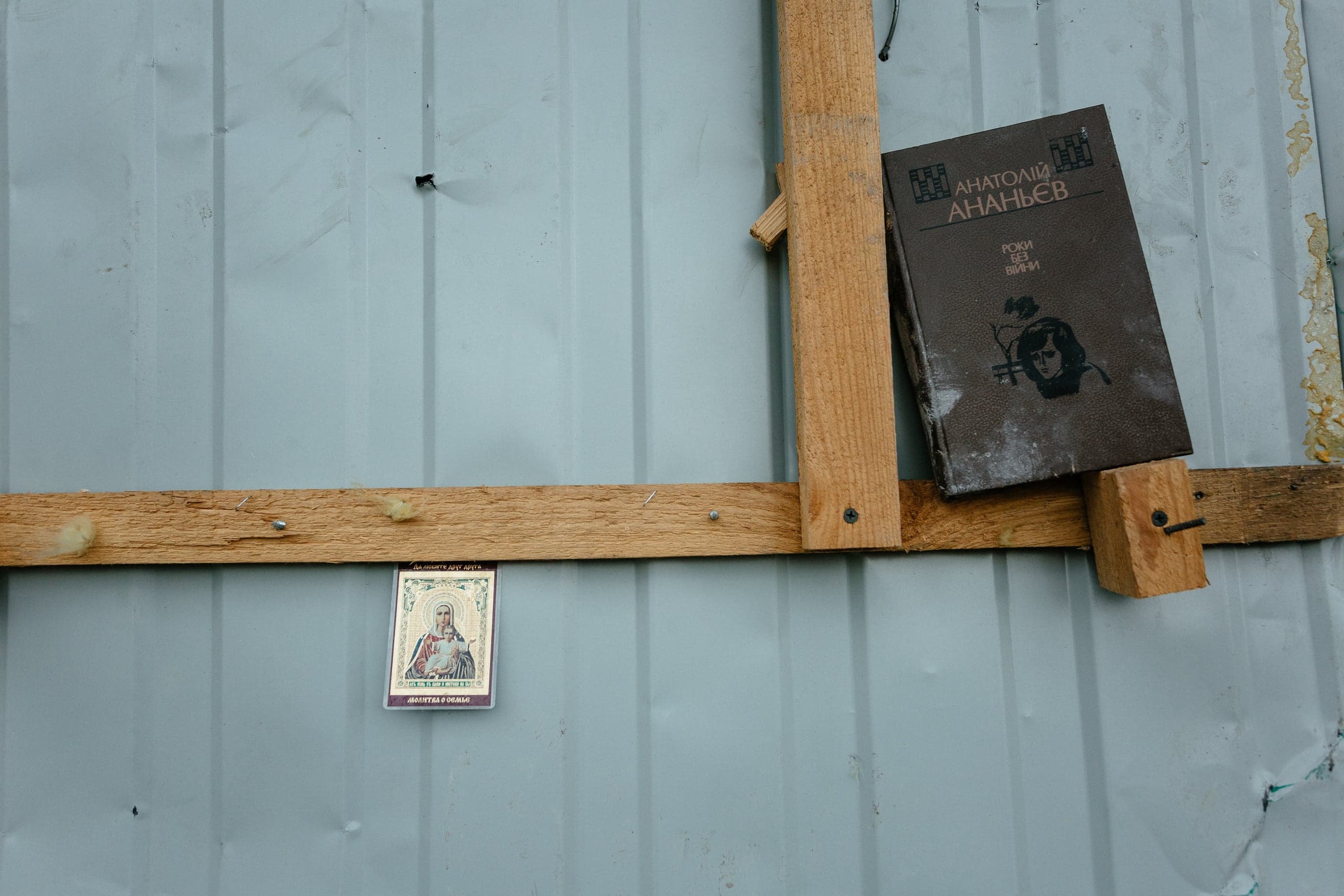 На металлической стене прикреплены деревянные рейки; на них лежат книга "Анатолій АНАНЬЄВ. РОКИ БЕЗ ВІЙНИ" и небольшая икона.
