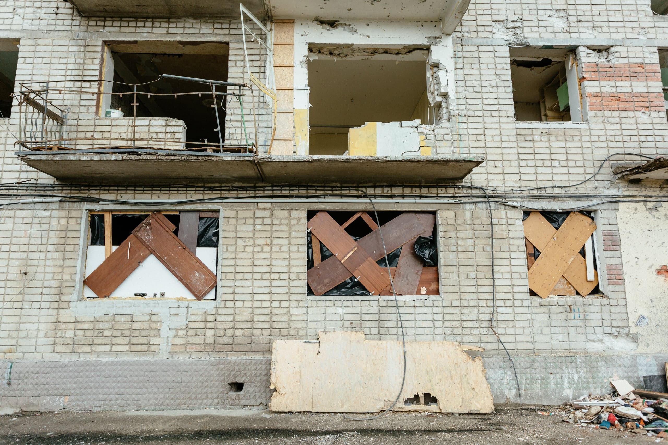Поврежденный кирпичный фасад жилого дома: верхние оконные проемы разрушены, нижние заколочены досками крест-накрест. У основания здания виден строительный мусор.