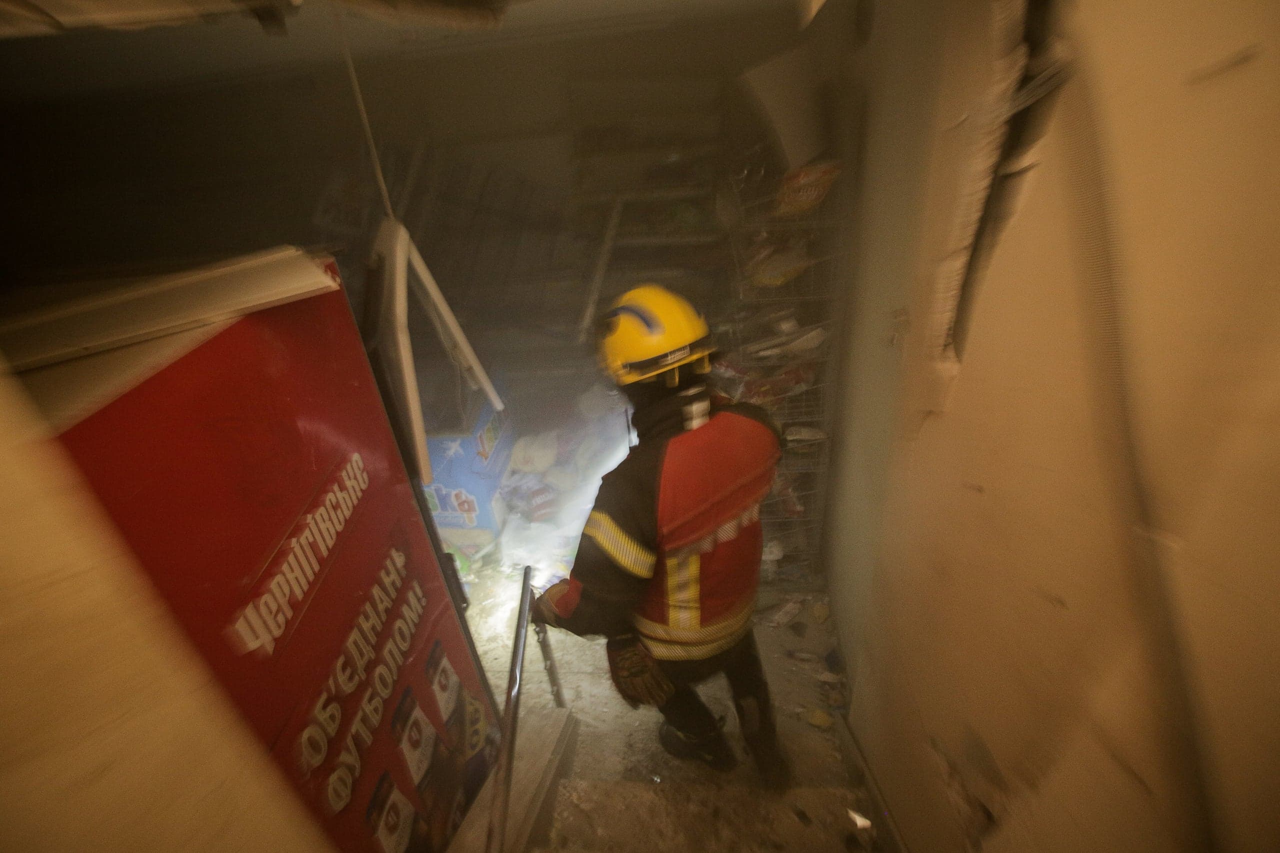Спасатель в желтой каске в задымленном помещении. Слева красный плакат: "Чернігівське", "ОБ'ЄДНАНІ ФУТБОЛОМ!".