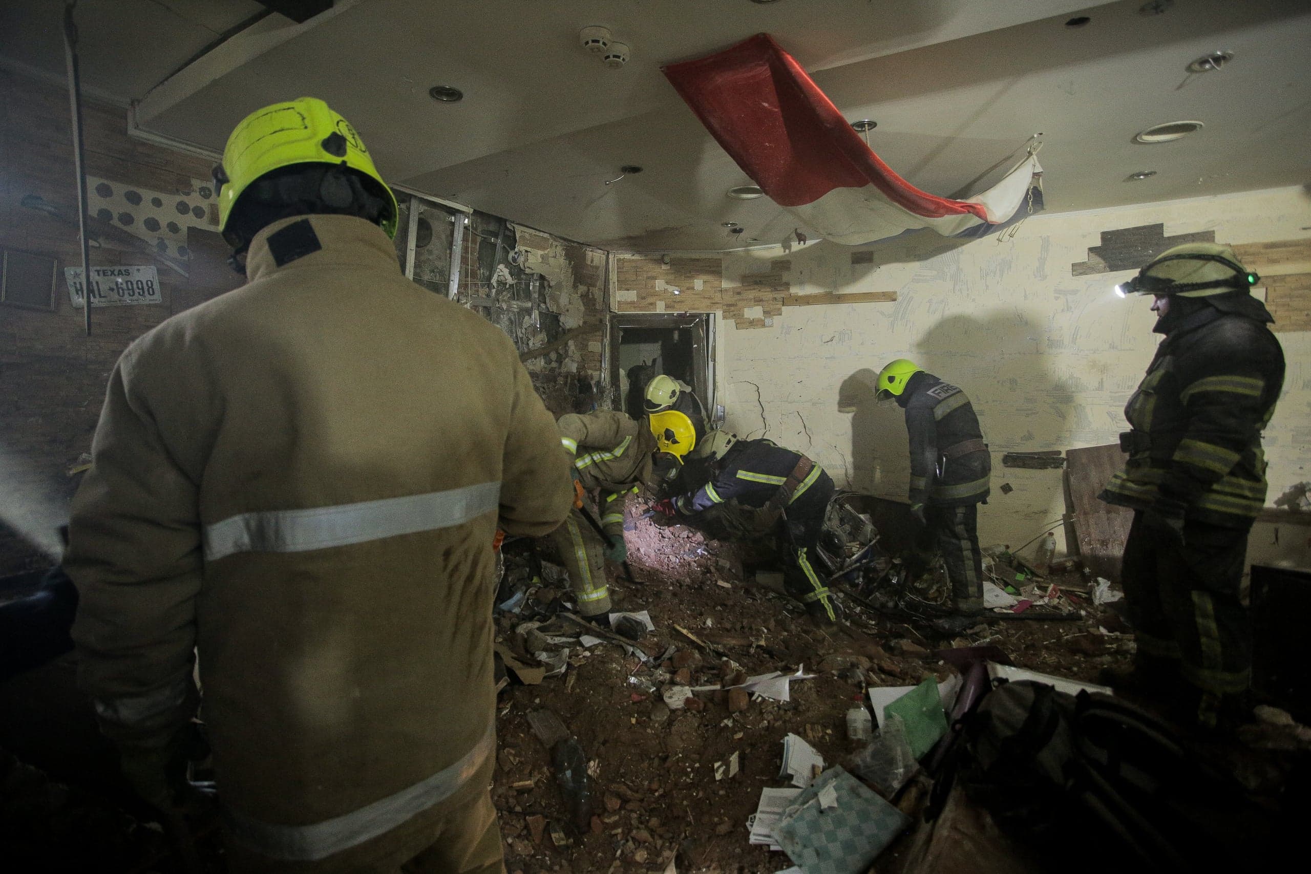 Четверо спасателей работают среди завалов в разрушенном помещении. На стене виден номерной знак "TEXAS HAL-6998".