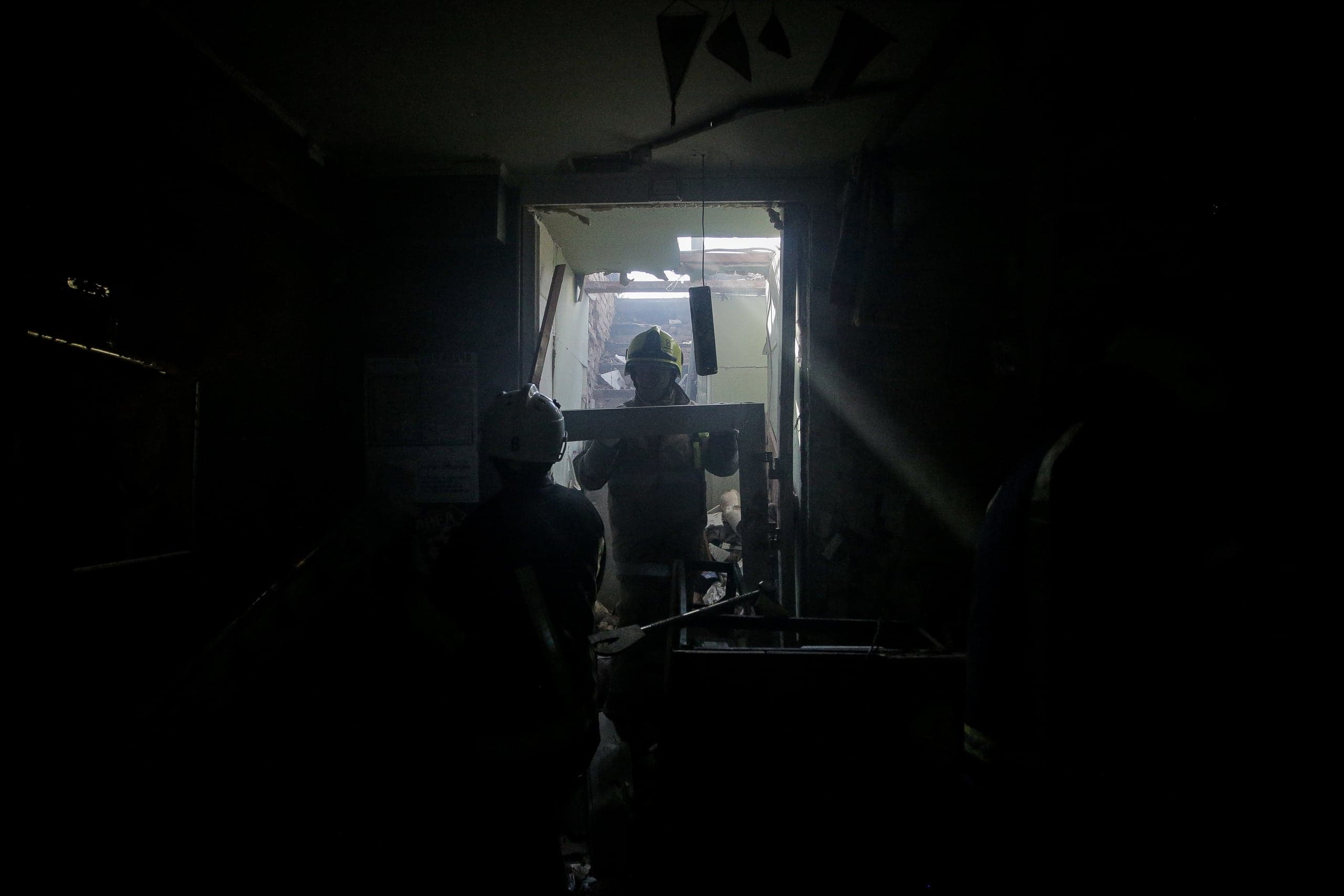 Двое спасателей в темном помещении. Один, освещенный сзади, несет конструкцию через дверной проем.