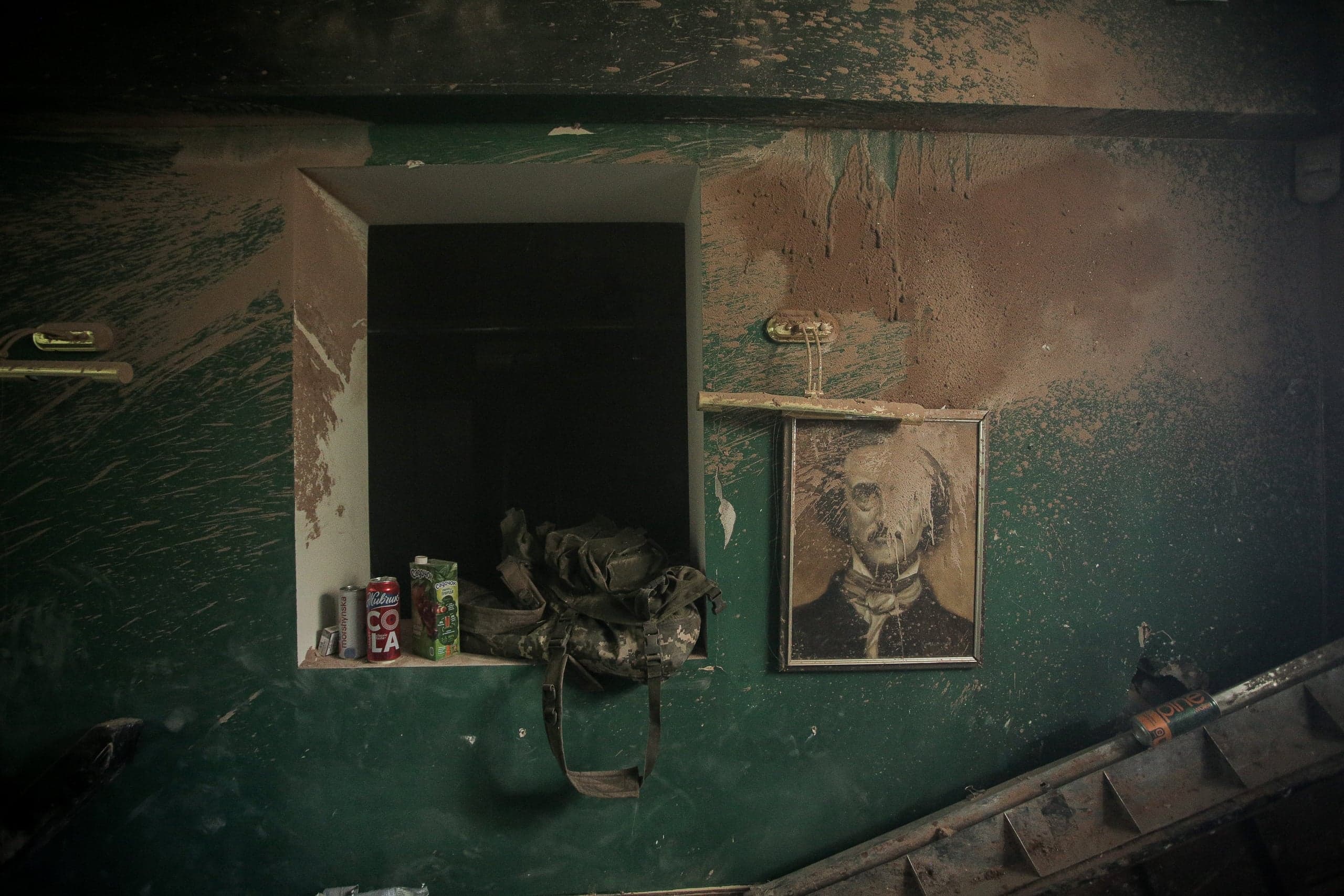 Загрязненный интерьер: зеленая стена, покрытая брызгами грязи, с нишей. На полке банка "Coca Cola" и камуфляжный рюкзак. Рядом висит испачканный портрет.