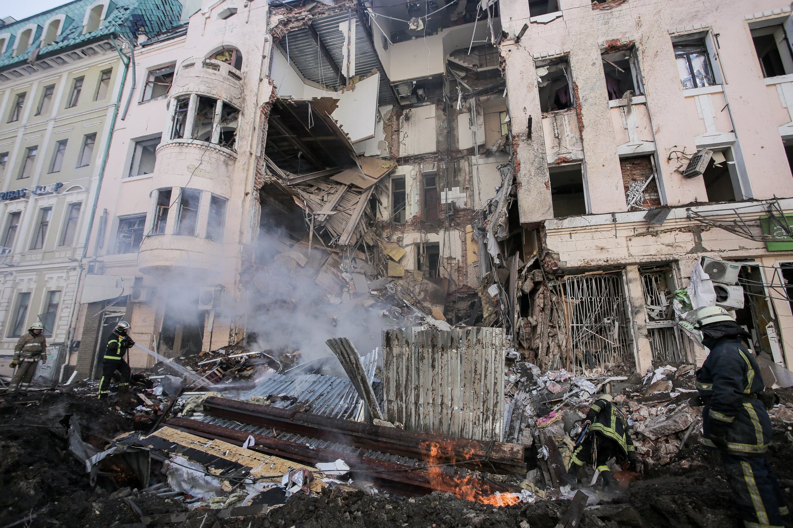 Разрушенный многоэтажный дом после удара; спасатели тушат пожар и задымленные завалы на переднем плане.