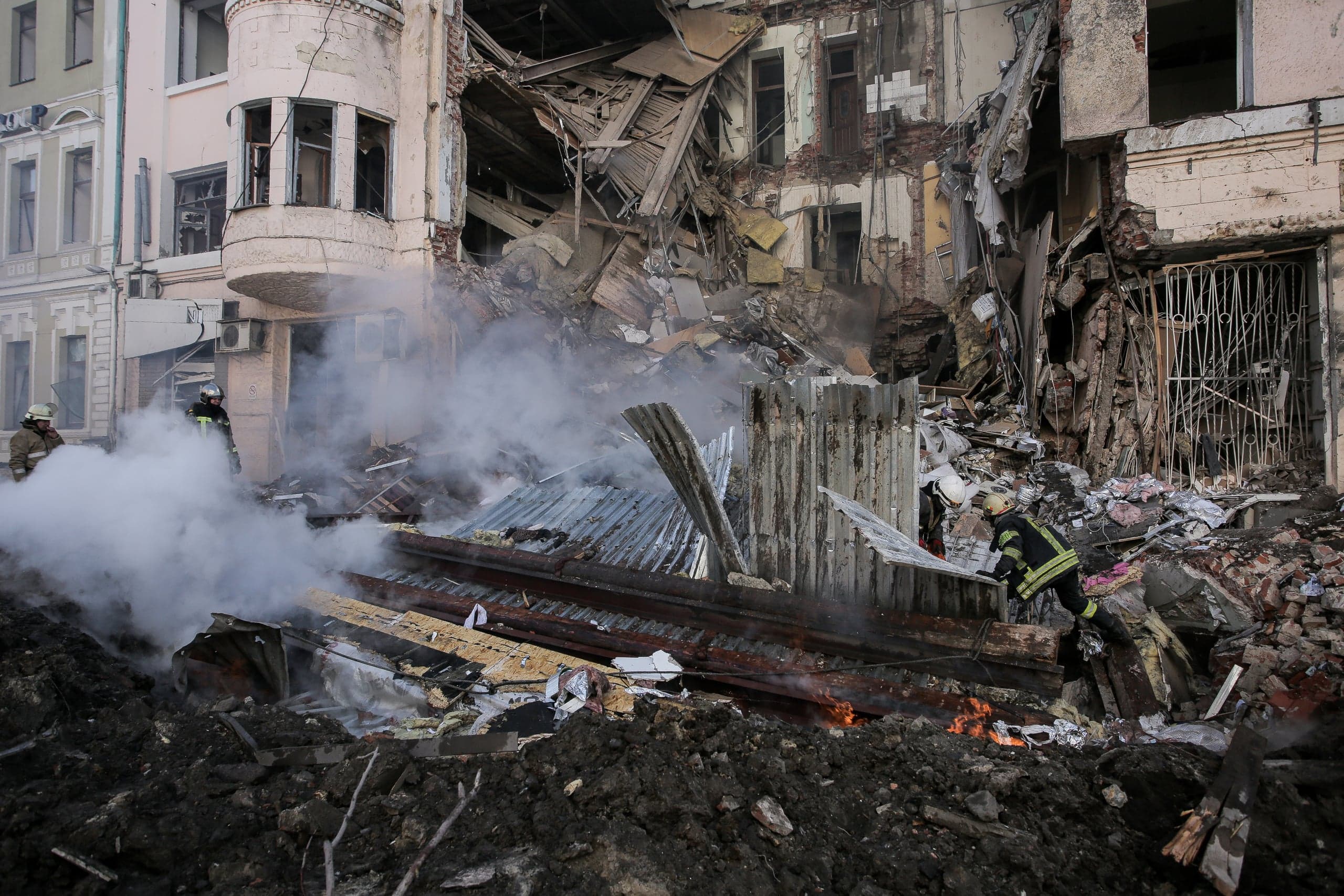 Разрушенное здание с рухнувшими перекрытиями; двое пожарных работают среди дымящихся обломков.