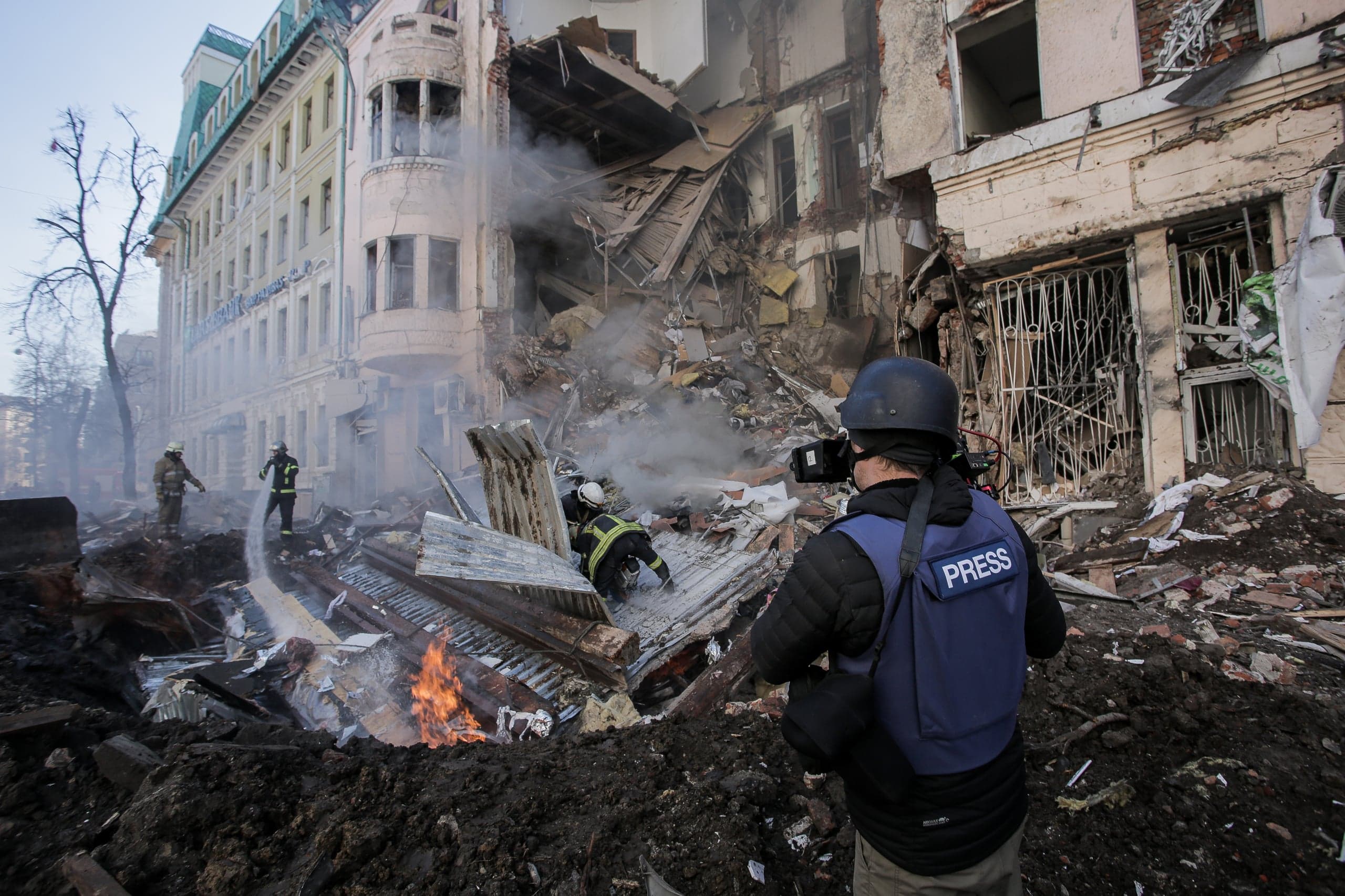 Журналист в жилете «PRESS» снимает сильно разрушенный взрывом дом; спасатели работают среди дымящихся обломков и огня.