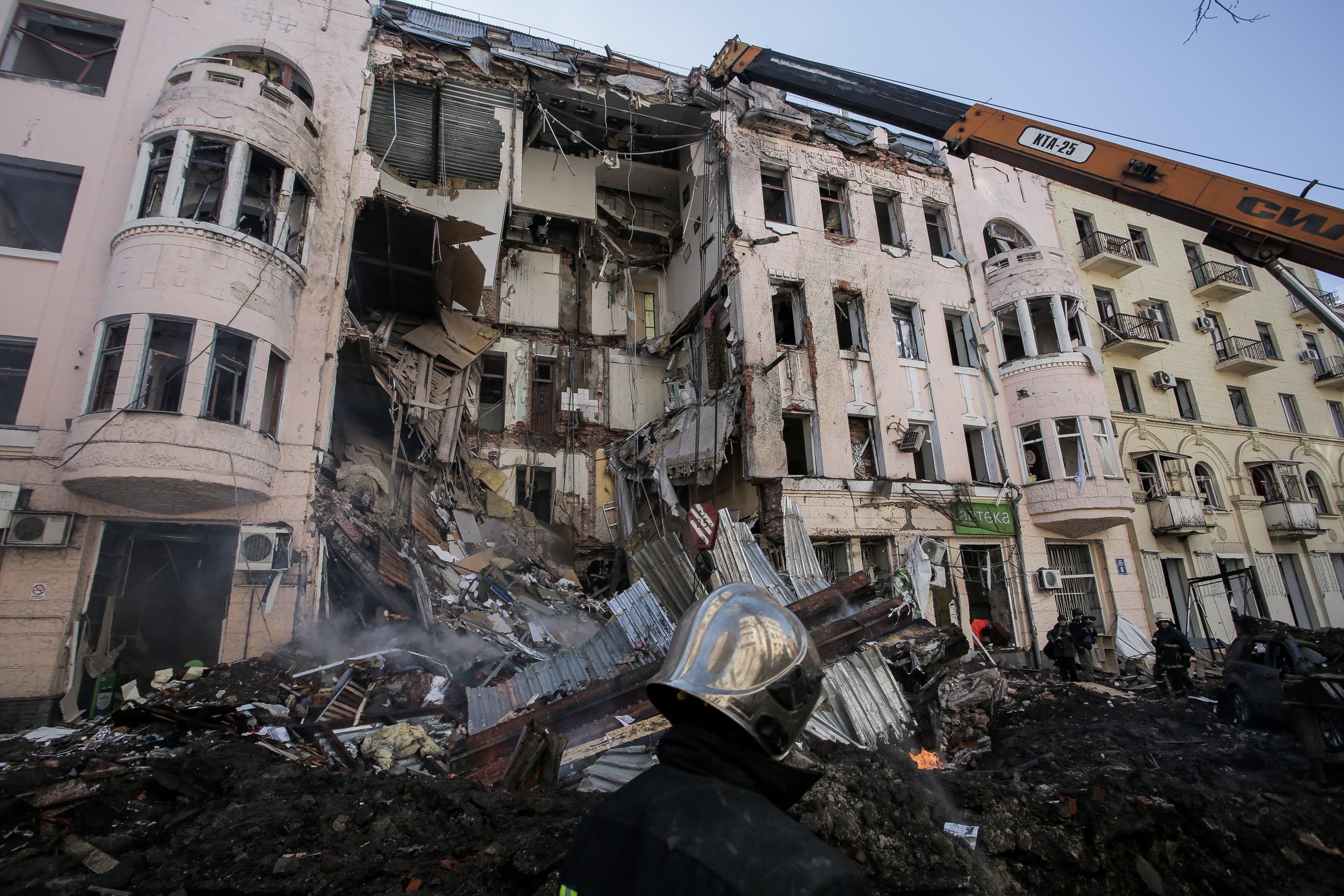 Разрушенное здание с обвалом в центре. Спасатель в каске стоит на фоне дымящихся завалов, над которыми видна стрела крана "КРА-25".