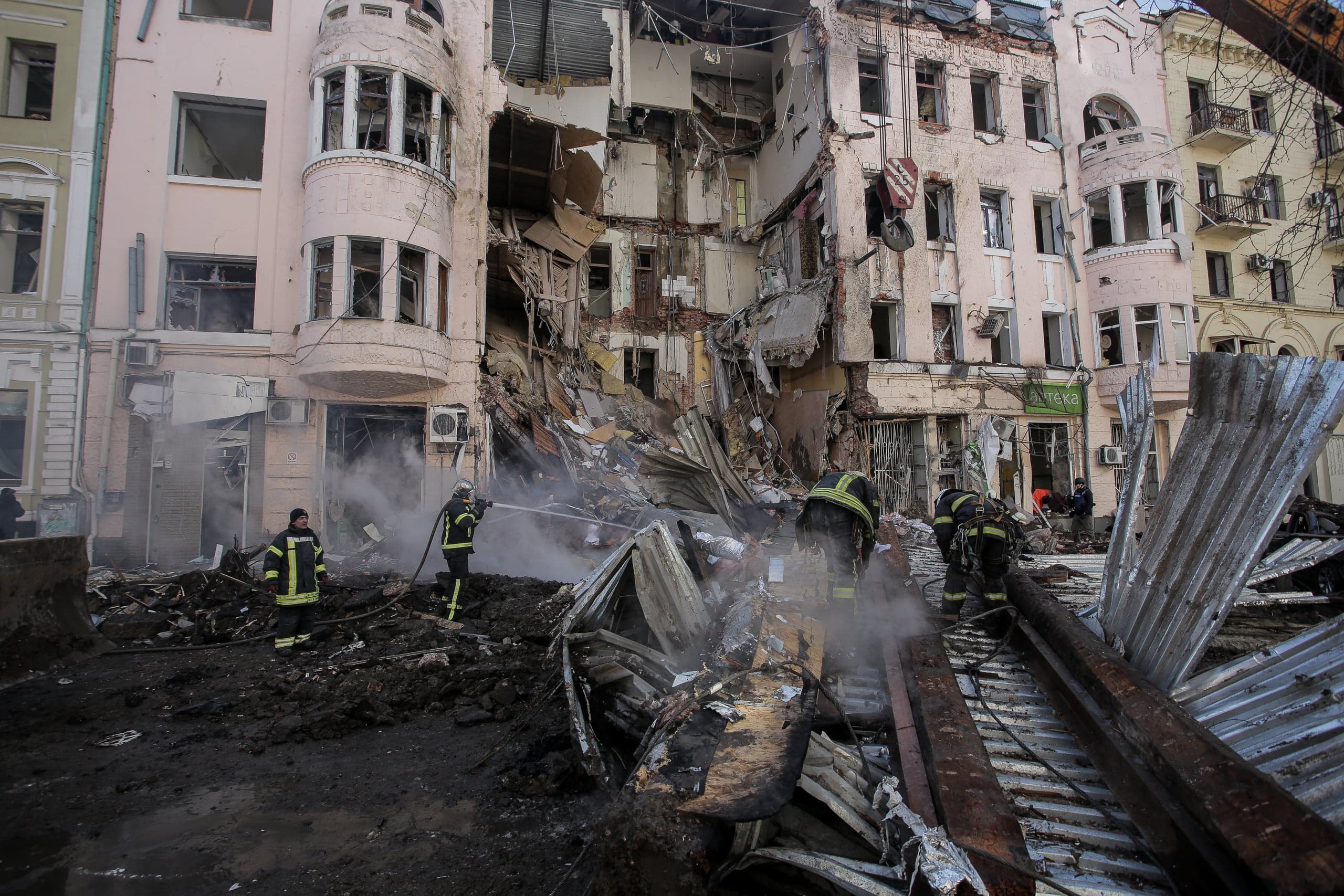 Пожарные работают среди руин частично обрушенного многоэтажного здания после обстрела. Обширные завалы, дым и строительный мусор. На уцелевшей части фасада видна вывеска "АПТЕКА".