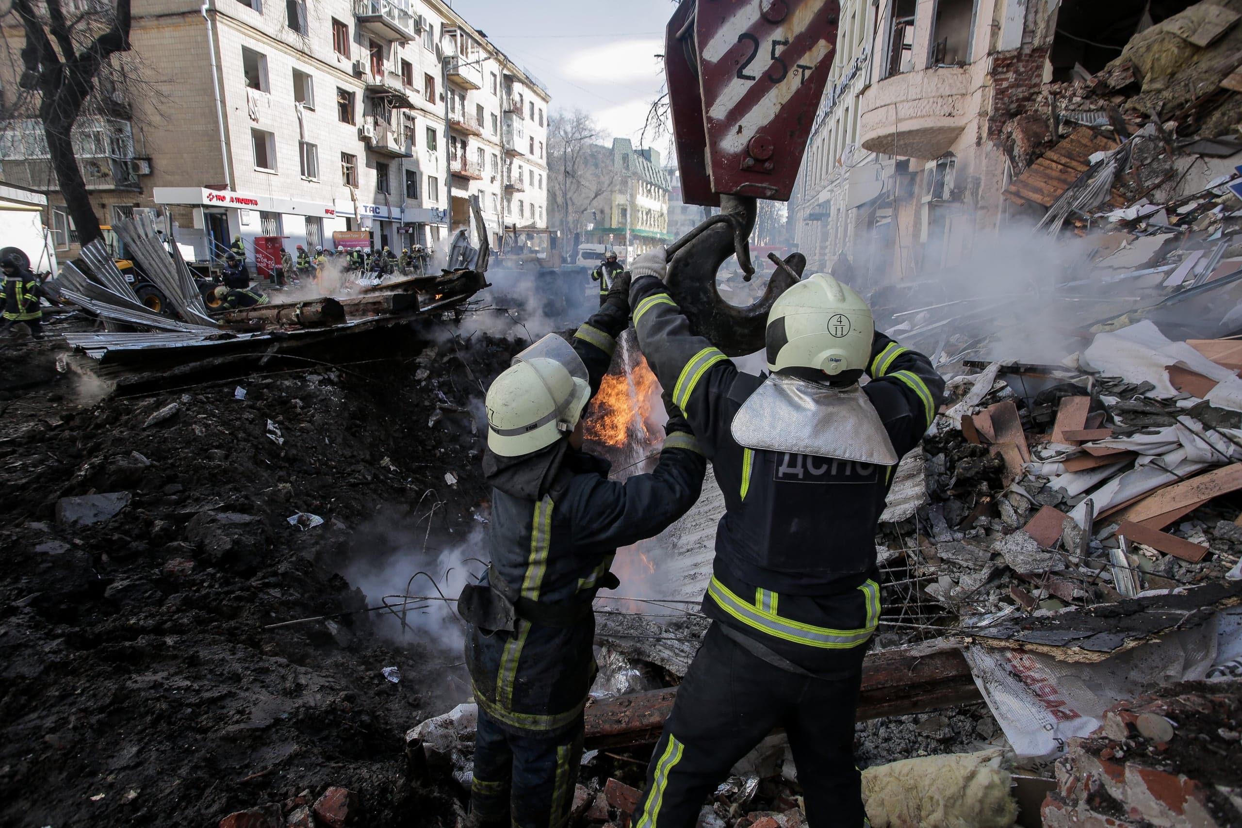 Два спасателя "ДСНС" работают у кранового крюка над очагом огня, на фоне руин и воронки в городском районе.