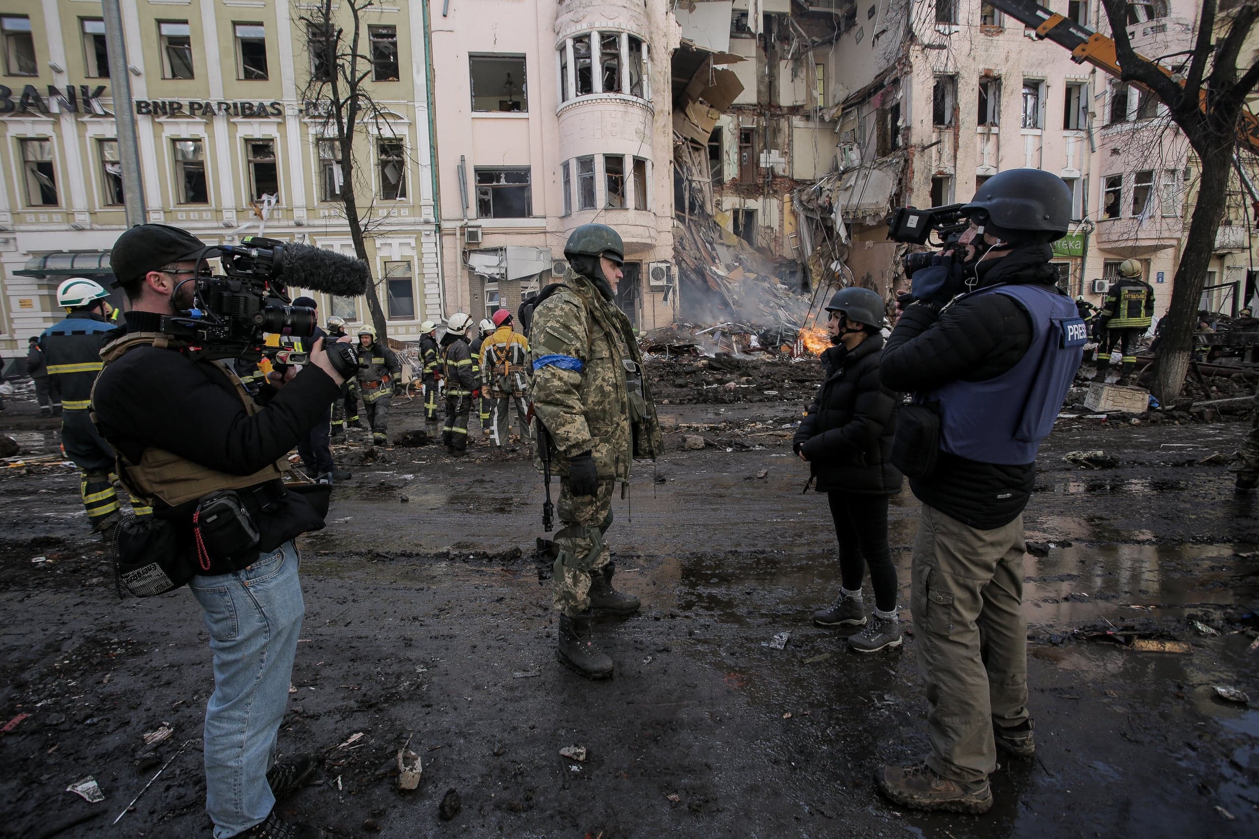 Военнослужащие, журналисты в жилетах "PRESS" с камерами и спасатели на фоне частично разрушенного здания с вывеской "BNP PARIBAS" после обстрела.