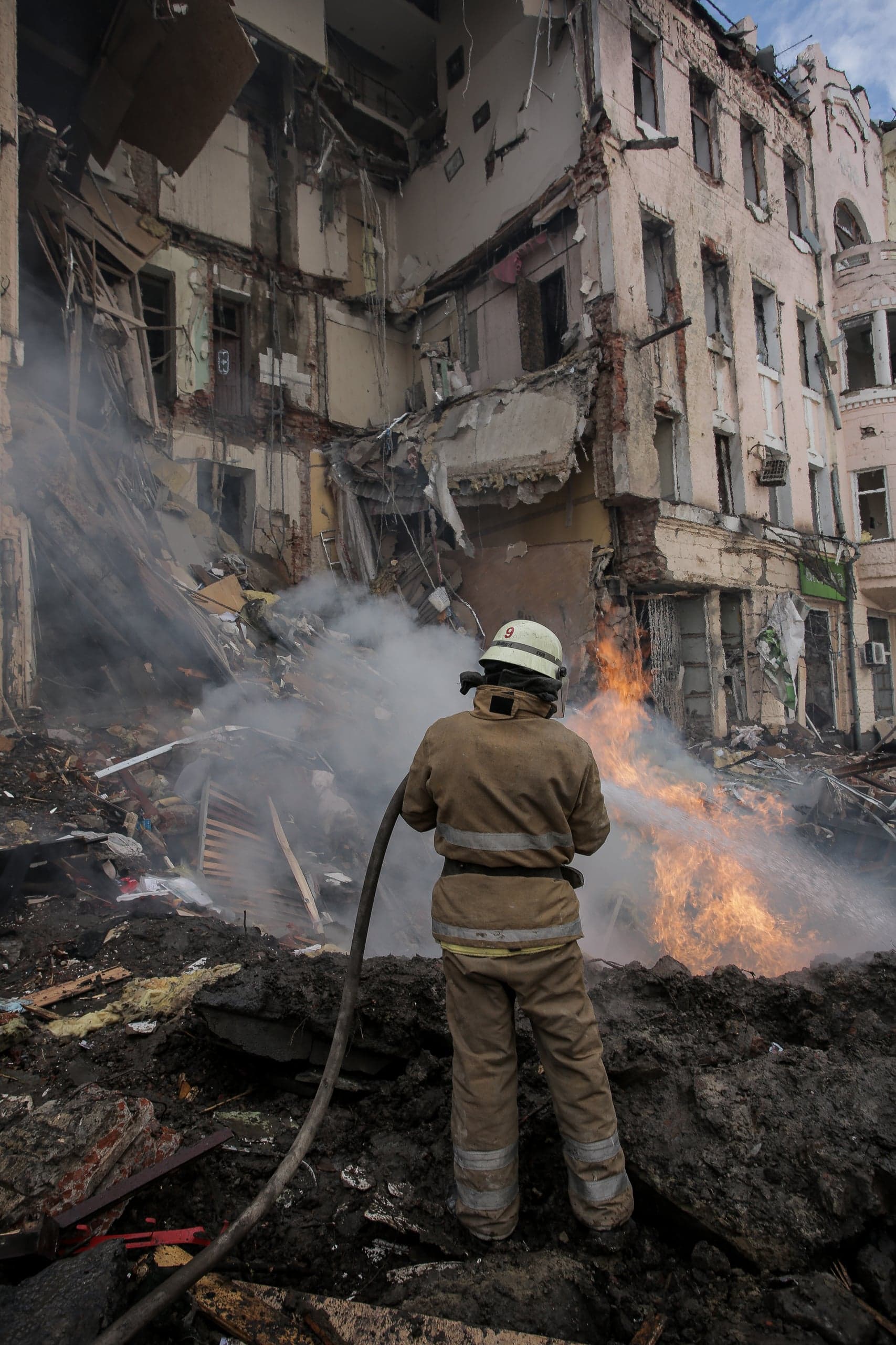 Пожарный тушит сильный огонь на фоне разрушенного многоэтажного здания, среди дыма и обломков.