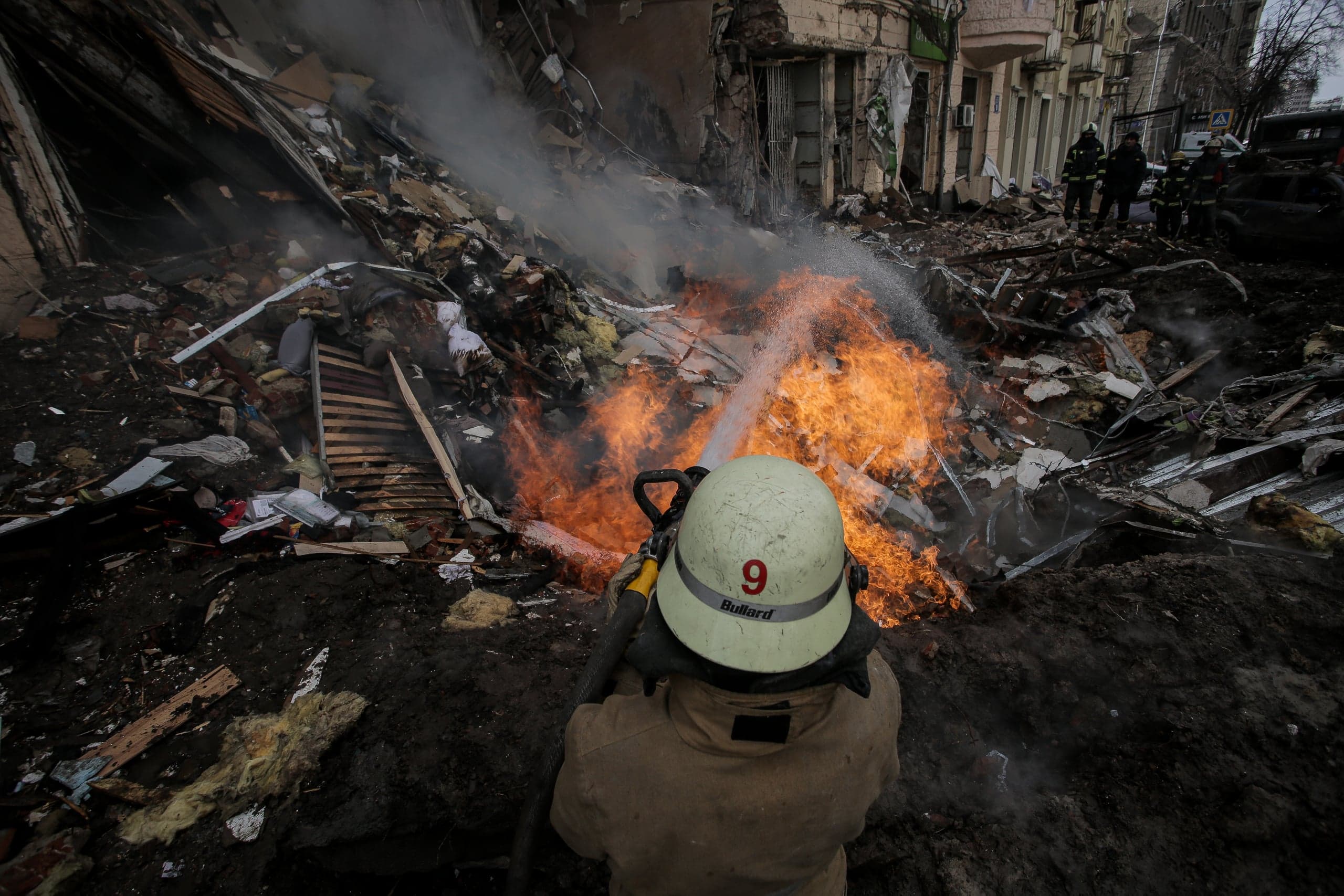Пожарный тушит сильный огонь среди руин разрушенного здания; на шлеме видны номер "9" и слово "Bullard".