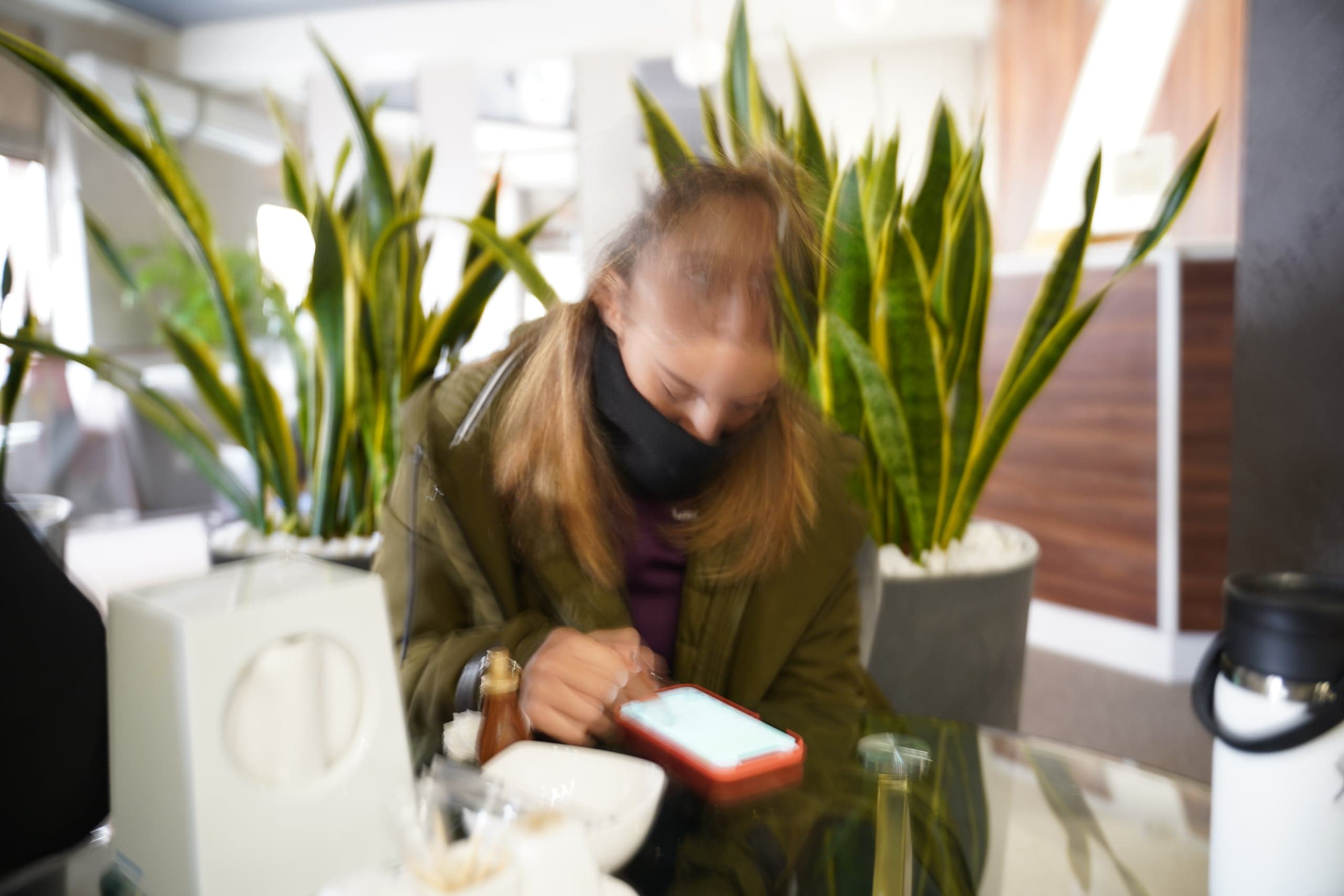 Размытый кадр: человек в зеленой куртке и черной маске сидит за столом и смотрит в смартфон. На заднем плане — высокие растения сансевиерии в горшках.