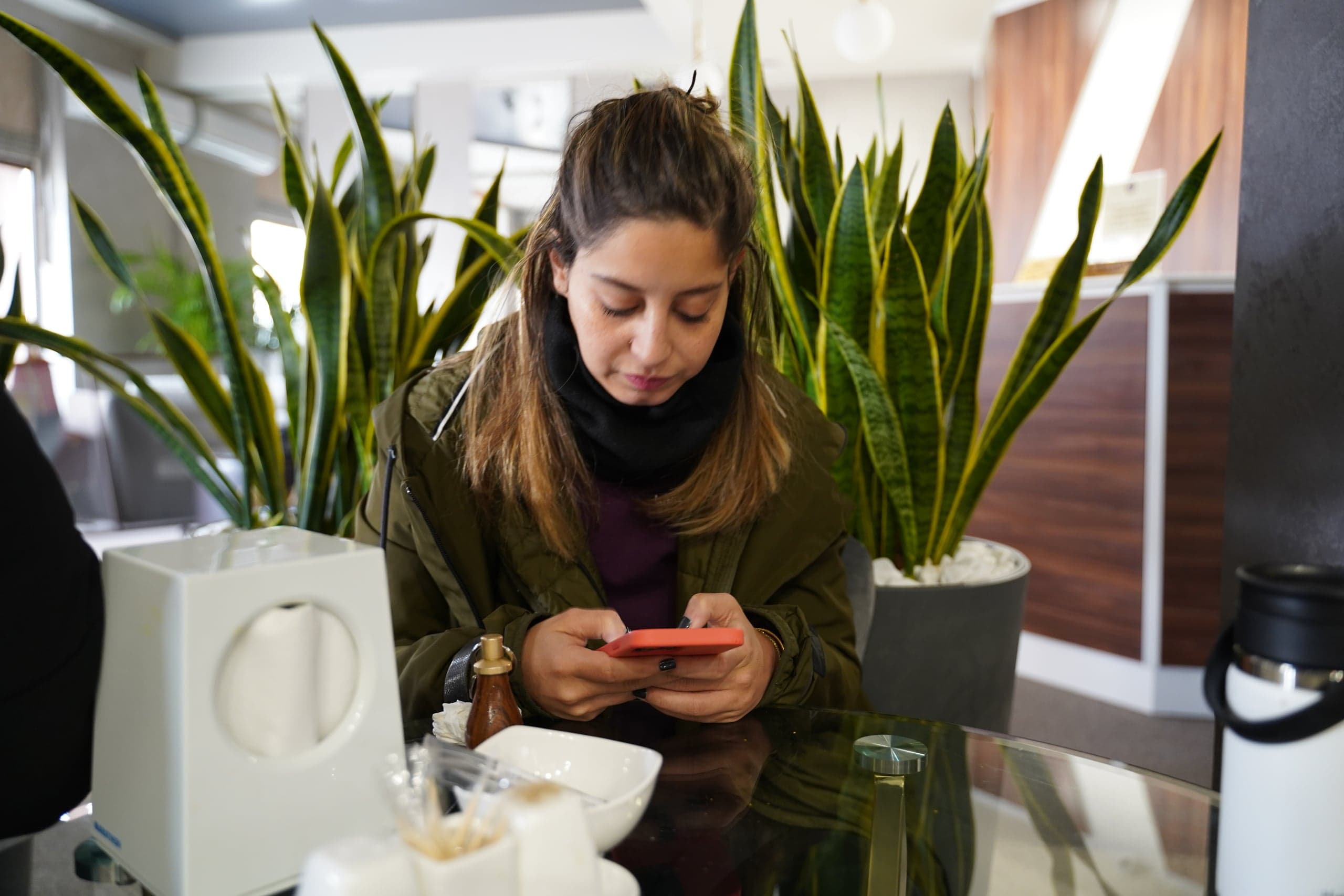 Женщина в куртке цвета хаки сидит за стеклянным столом, склонившись над оранжевым смартфоном, на фоне высоких горшечных растений (сансевиерий).