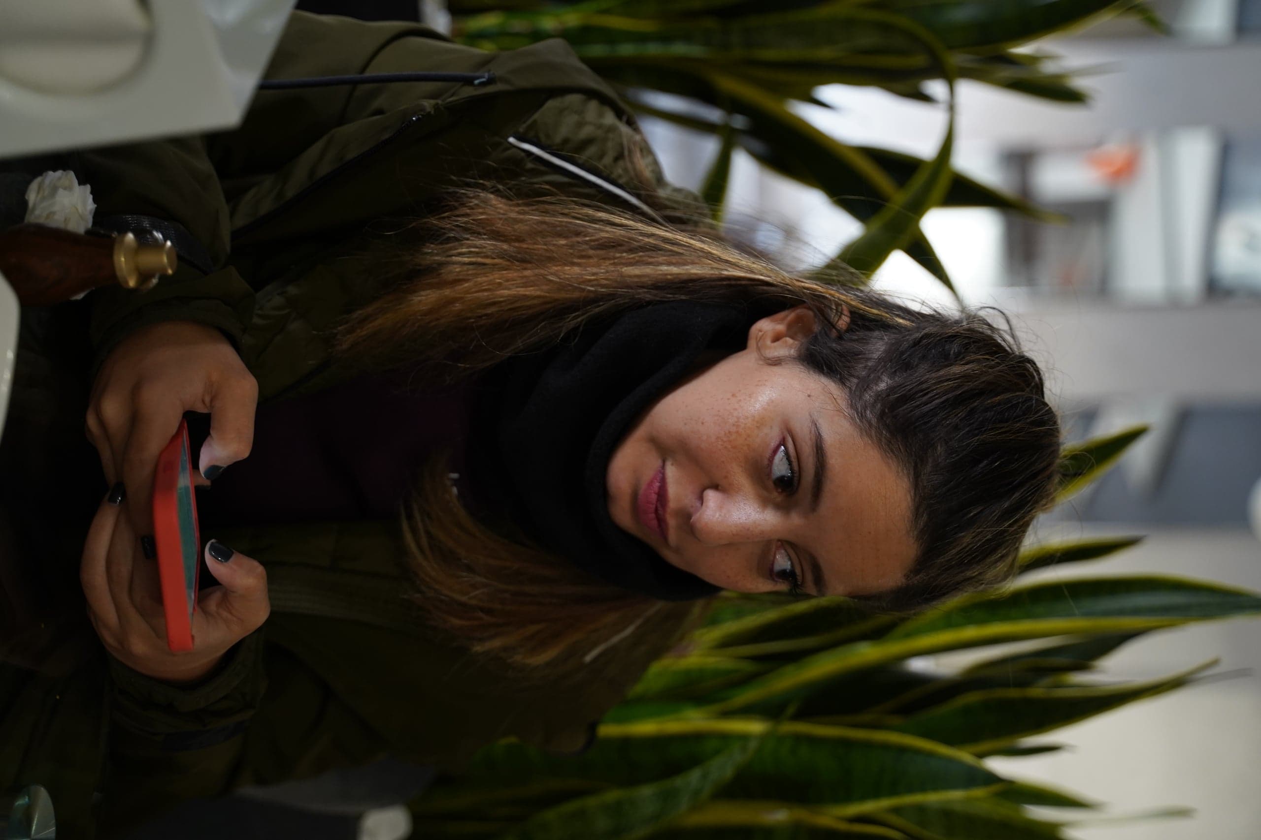 Крупный план женщины в оливковой куртке, смотрящей в сторону и держащей смартфон, на фоне высоких зеленых растений (сансевиерий).