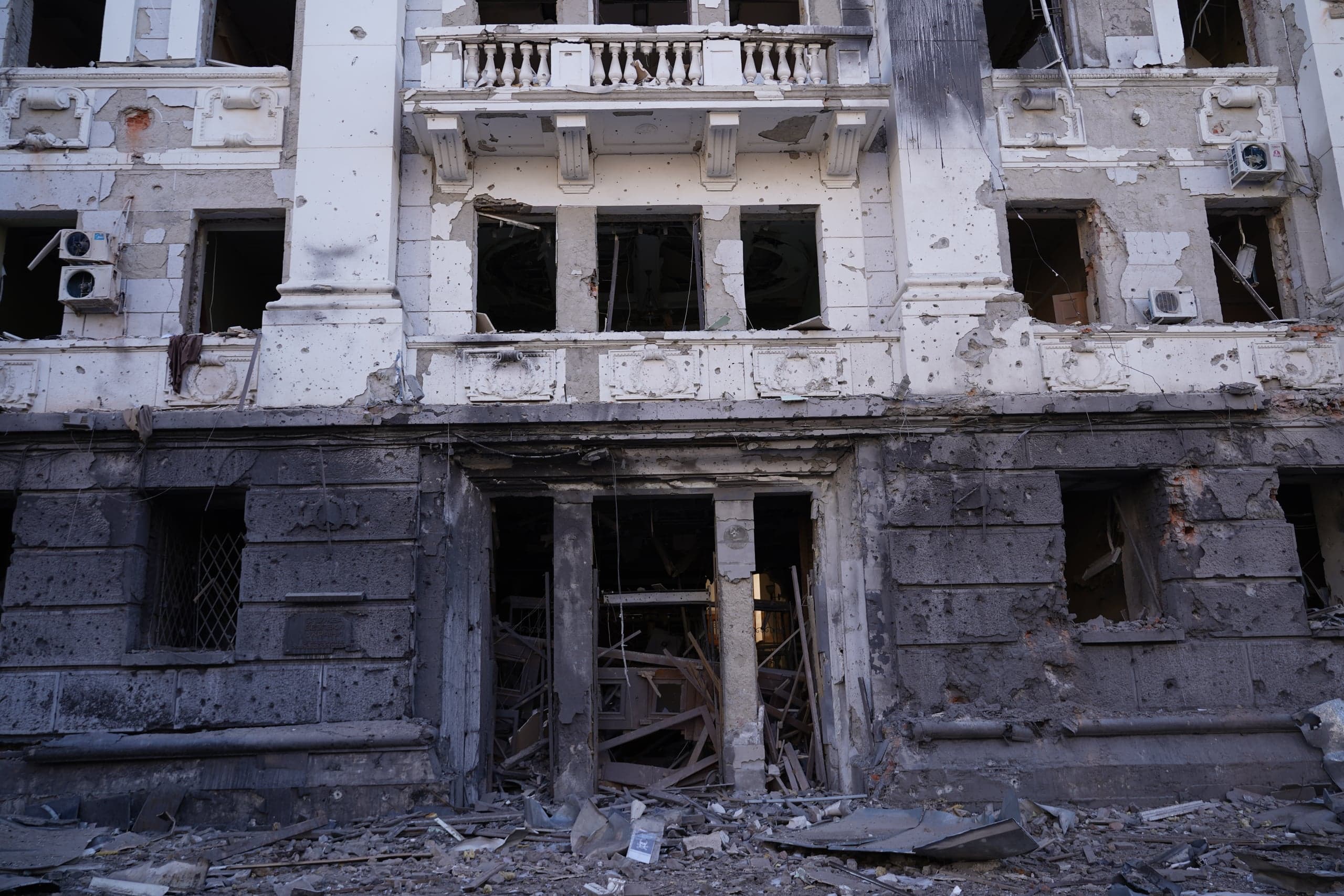 Разрушенный фасад здания, верхняя часть белая, нижняя тёмная. Поверхность испещрена следами от пуль и осколков, вход и окна выбиты, а на земле лежит груда обломков.