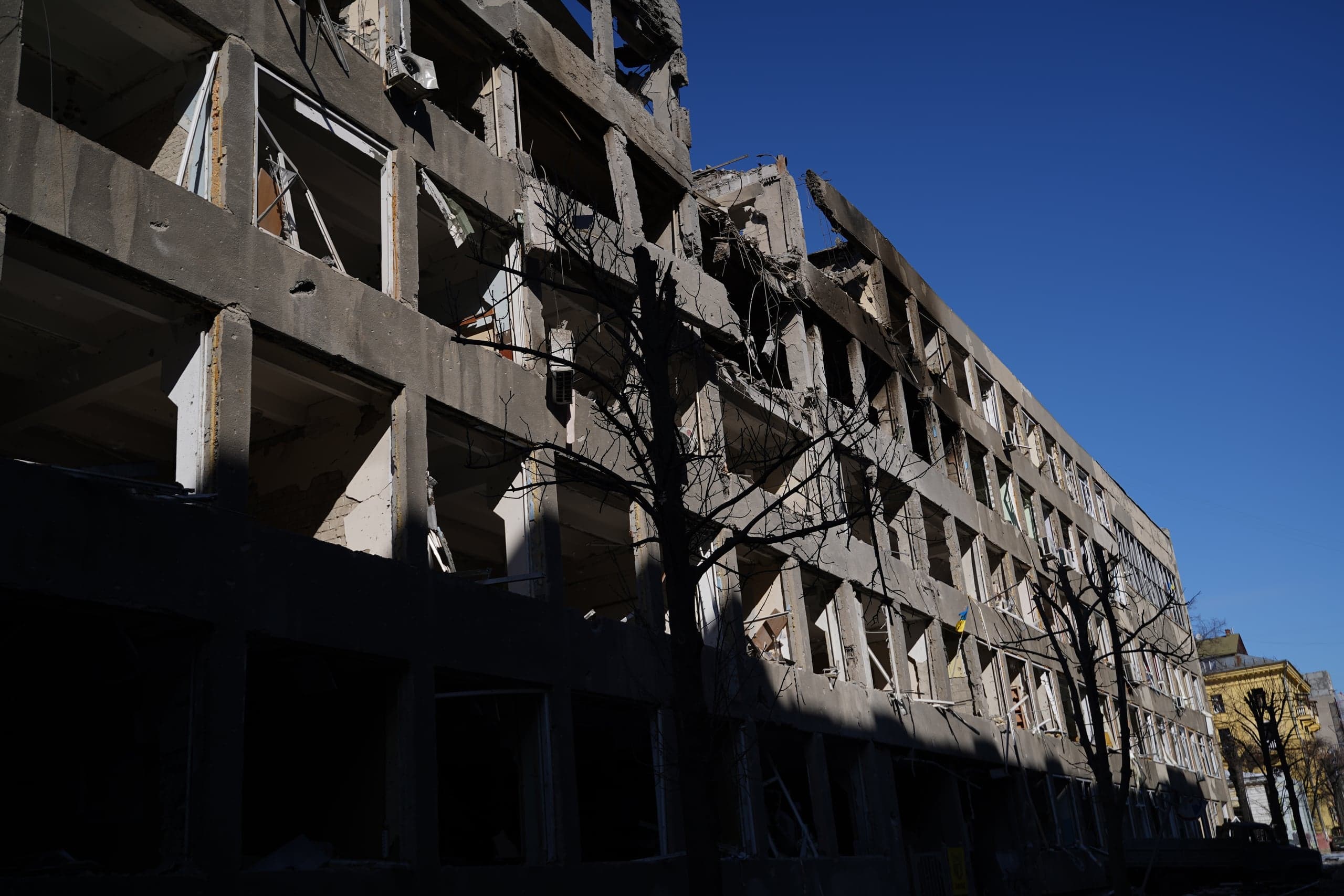 Фасад сильно поврежденного бетонного многоэтажного здания с выбитыми окнами на фоне ярко-голубого неба. Левая часть здания в глубокой тени. На одном из окон виден маленький флаг Украины.