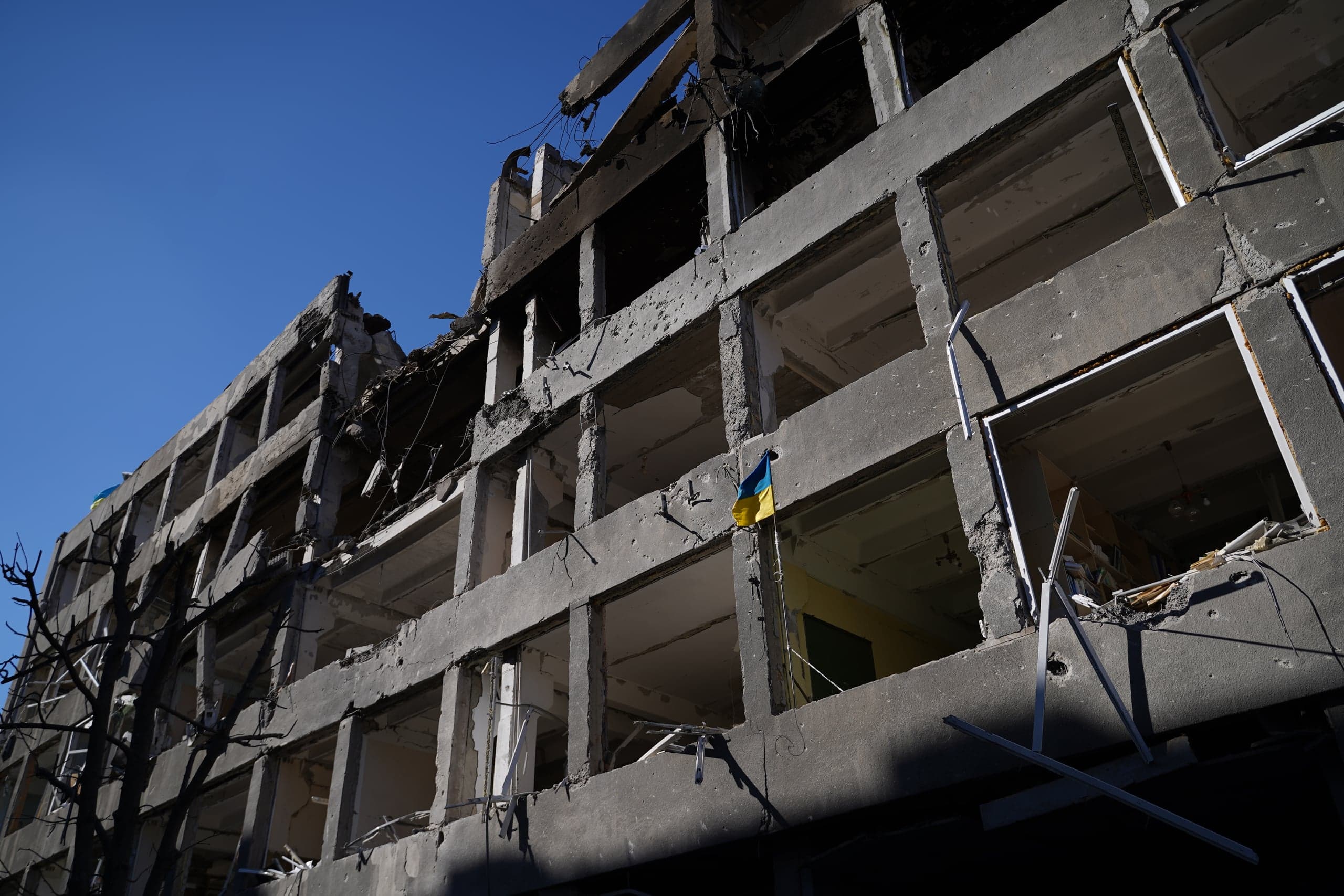 Фасад сильно разрушенного бетонного здания с пустыми оконными проемами и следами повреждений. На одной из балок висит маленький флаг Украины.