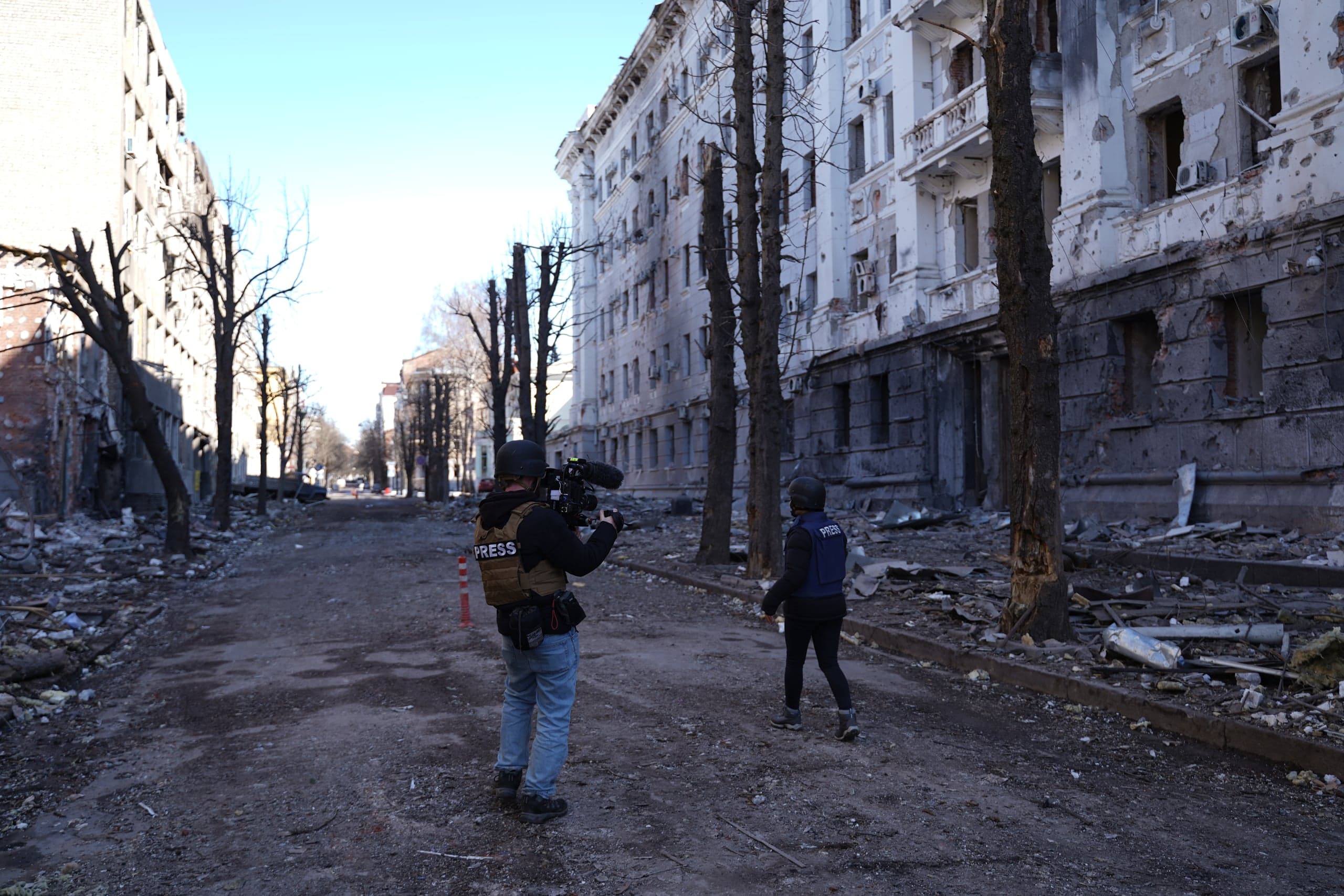 Два журналиста в защитной экипировке "PRESS" снимают на видеокамеру разрушенный городской квартал. Улица усыпана обломками, а многоэтажные здания сильно повреждены обстрелами.