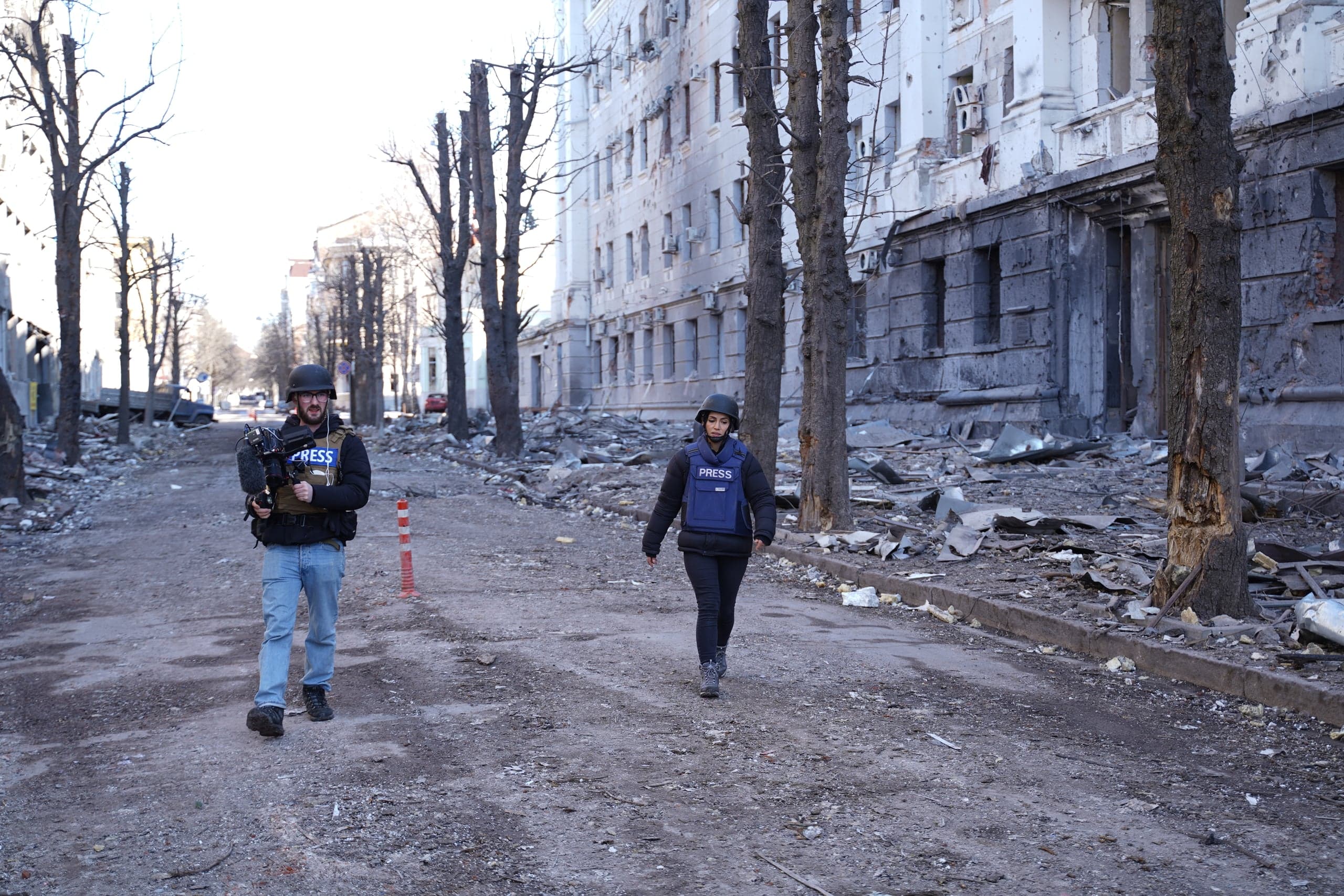 Двое журналистов (оператор с камерой и женщина) в шлемах и бронежилетах "PRESS" идут по разрушенной улице, усыпанной обломками, на фоне сильно поврежденных зданий.