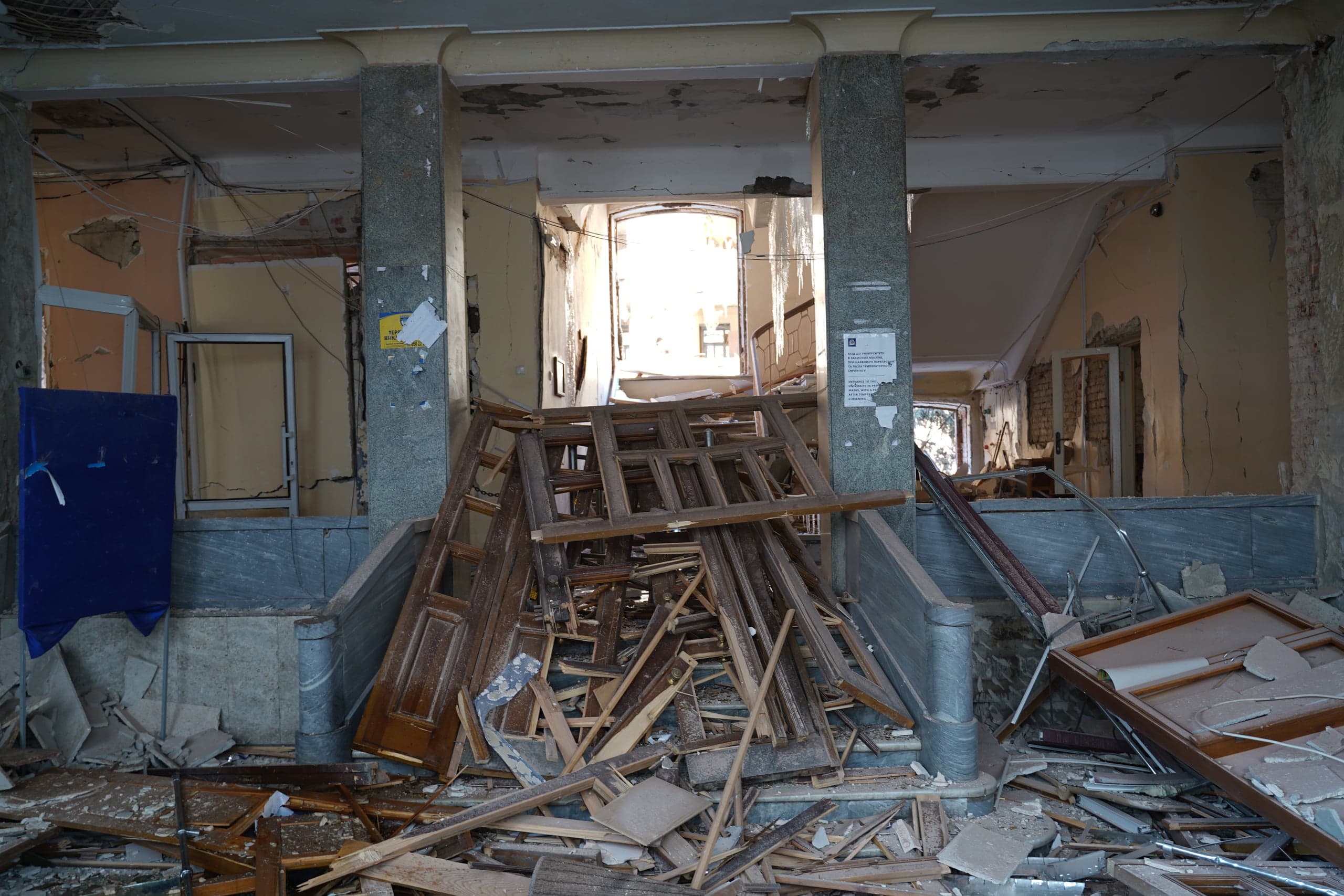 Сильно поврежденный интерьер вестибюля с двумя каменными колоннами, заваленный грудой обломков, разбитых деревянных оконных и дверных рам. Яркий свет виден в проеме на заднем плане.