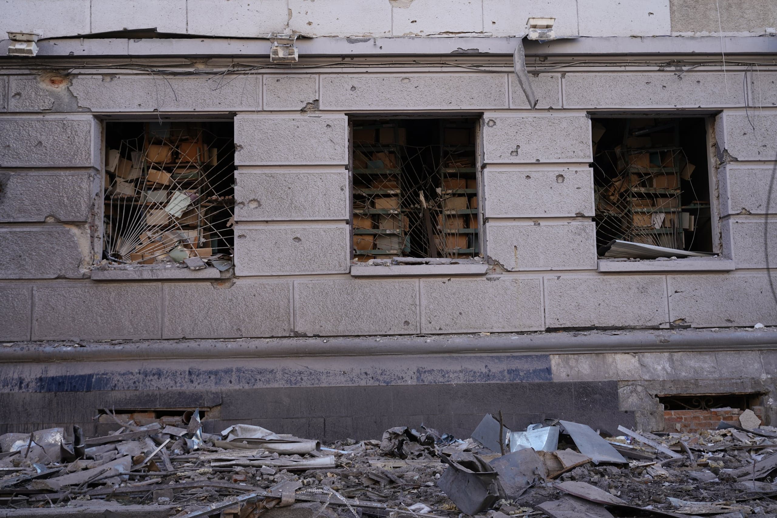Фасад поврежденного здания с тремя выбитыми окнами, за которыми видны изогнутые металлические решетки, и большой грудой строительного мусора на земле. На стене видны сколы и следы разрушений.