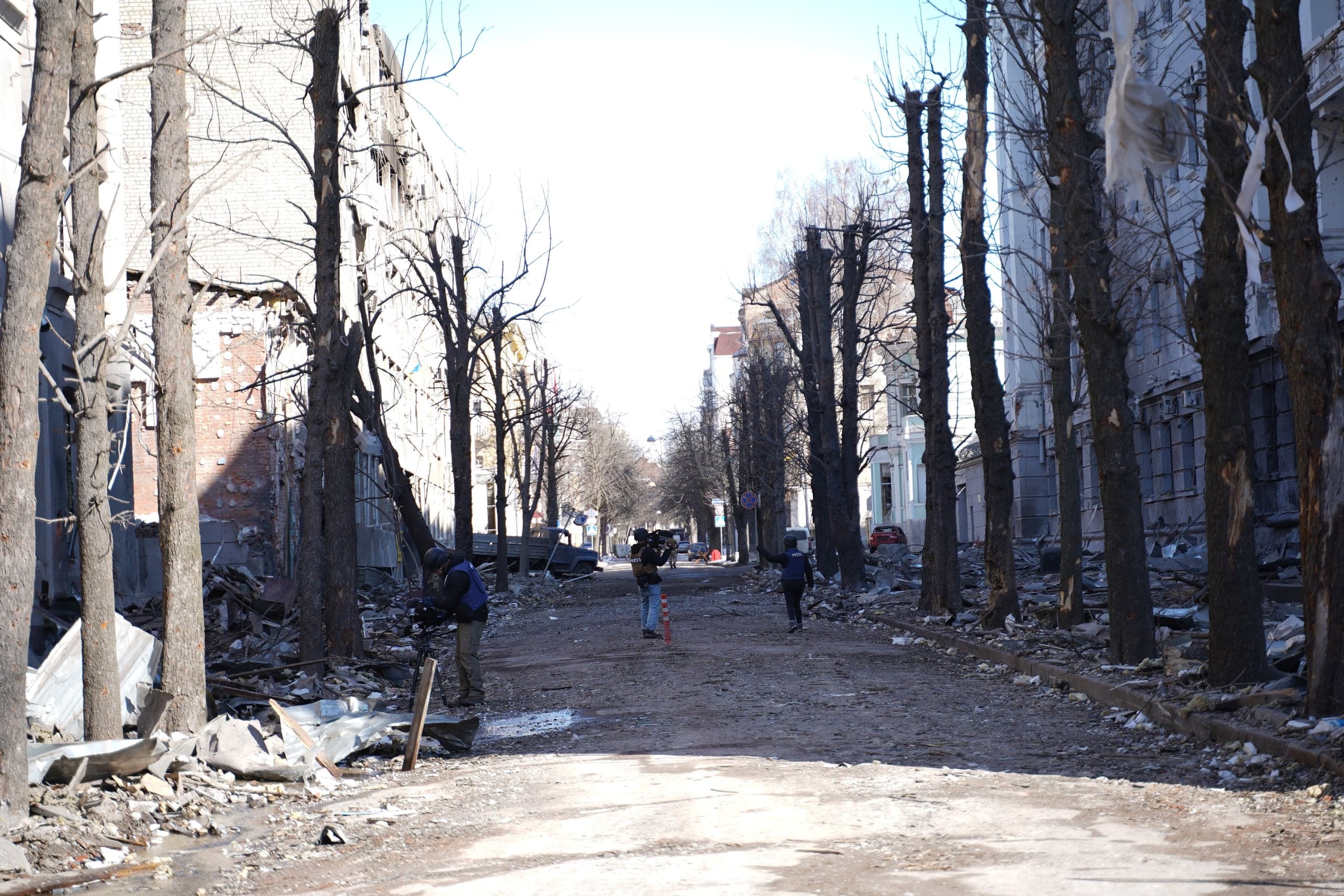 Улица, разрушенная обстрелами. По обе стороны видны сильно поврежденные здания и голые деревья. Трое людей, один из которых несет видеокамеру, идут по засыпанной обломками дороге.