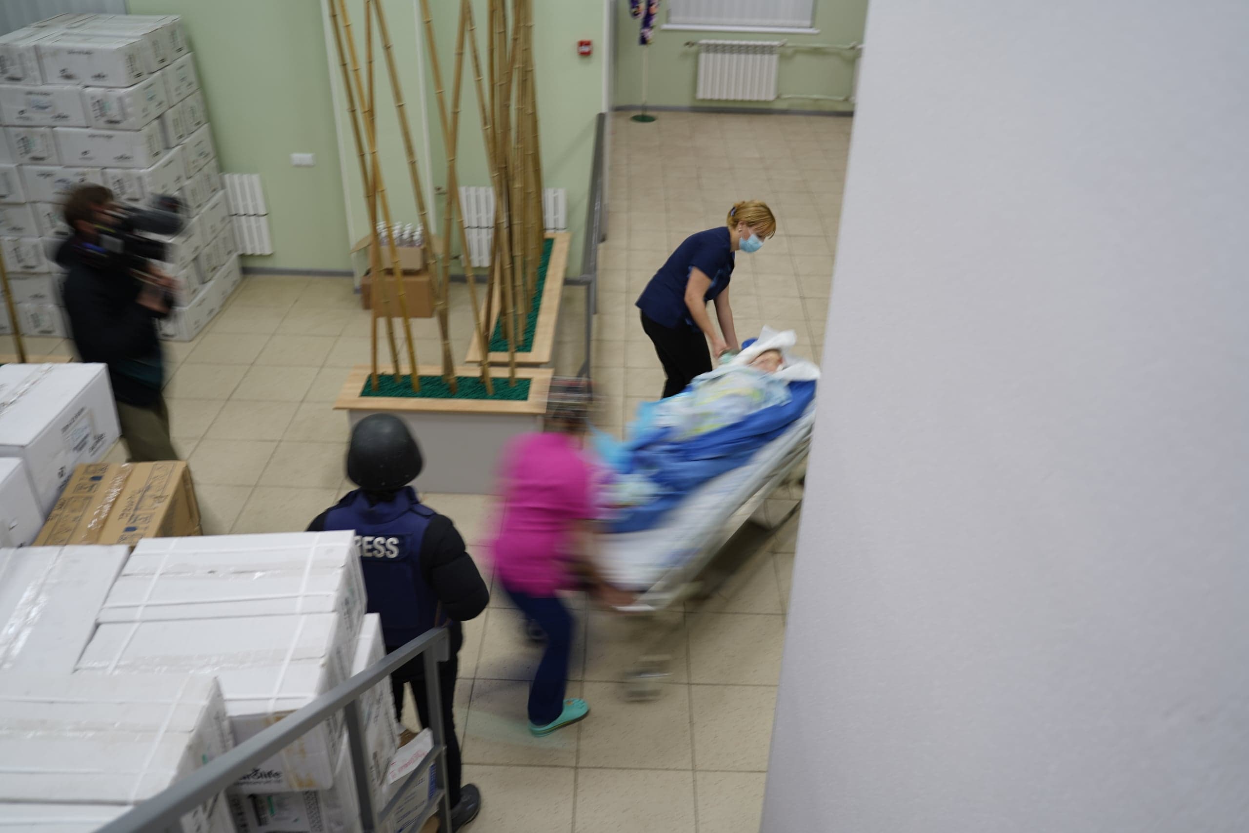 Больничный коридор, заставленный коробками. Медработники быстро везут пациента на каталке мимо журналистов с камерами и в жилетах «PRESS». Изображение с сильным размытием движения.