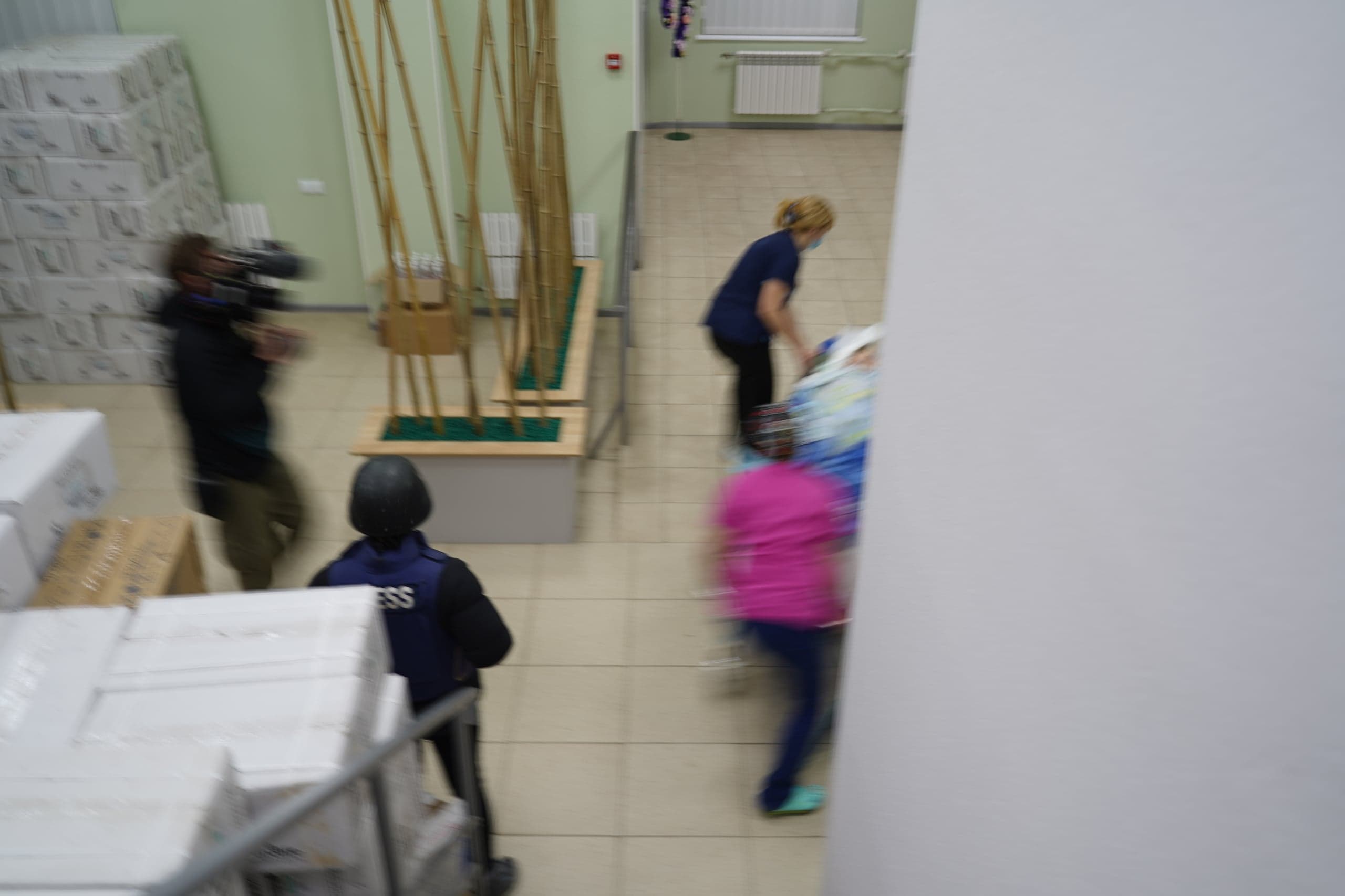 Размытый кадр: В помещении медики быстро везут каталку мимо высоких штабелей коробок. Слева видны оператор с камерой и человек в шлеме и жилете с надписью "PRESS".