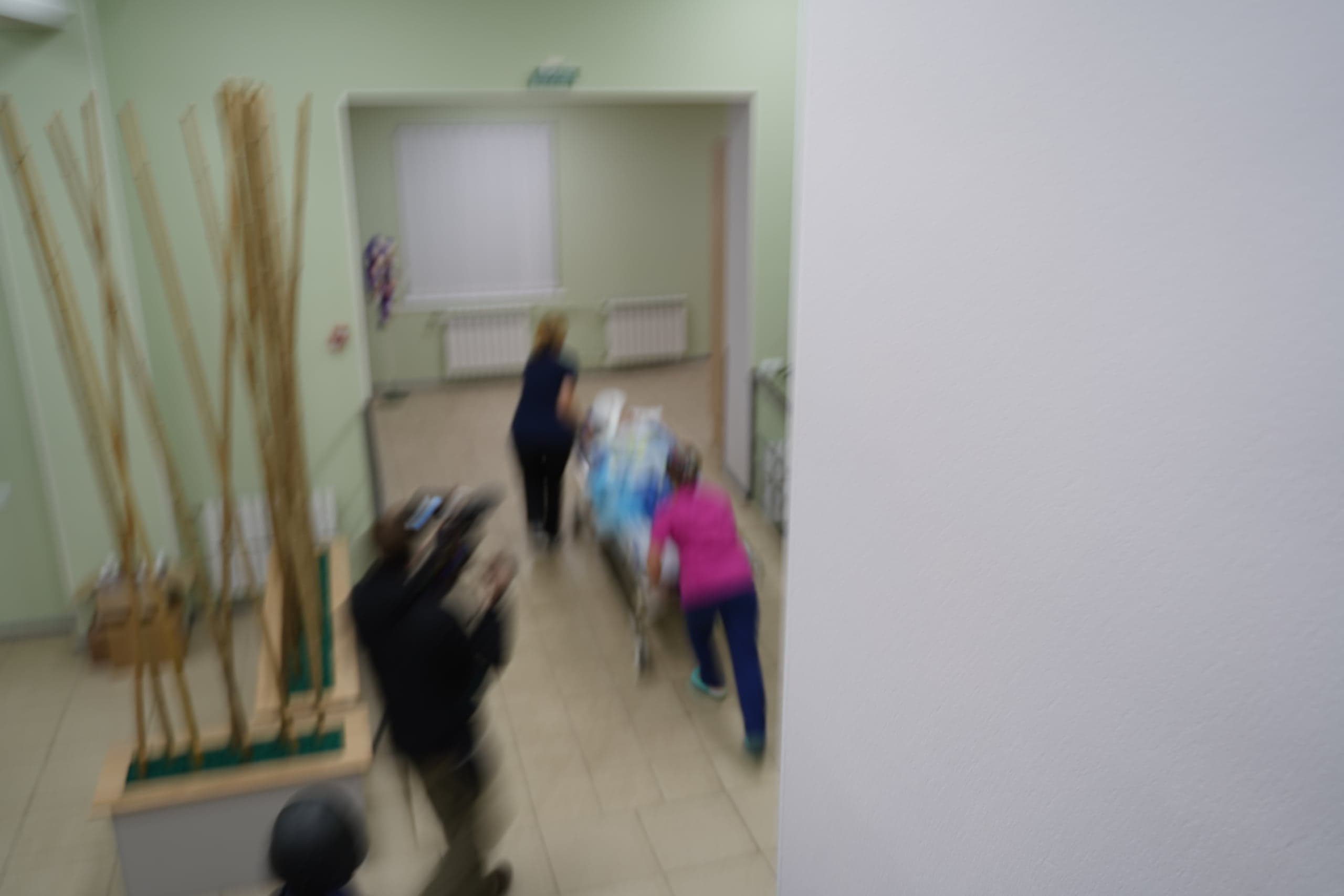 Сильно размытый кадр больничного коридора. Две фигуры, предположительно медперсонал, стремительно катят каталку с пациентом; слева виден человек с камерой.