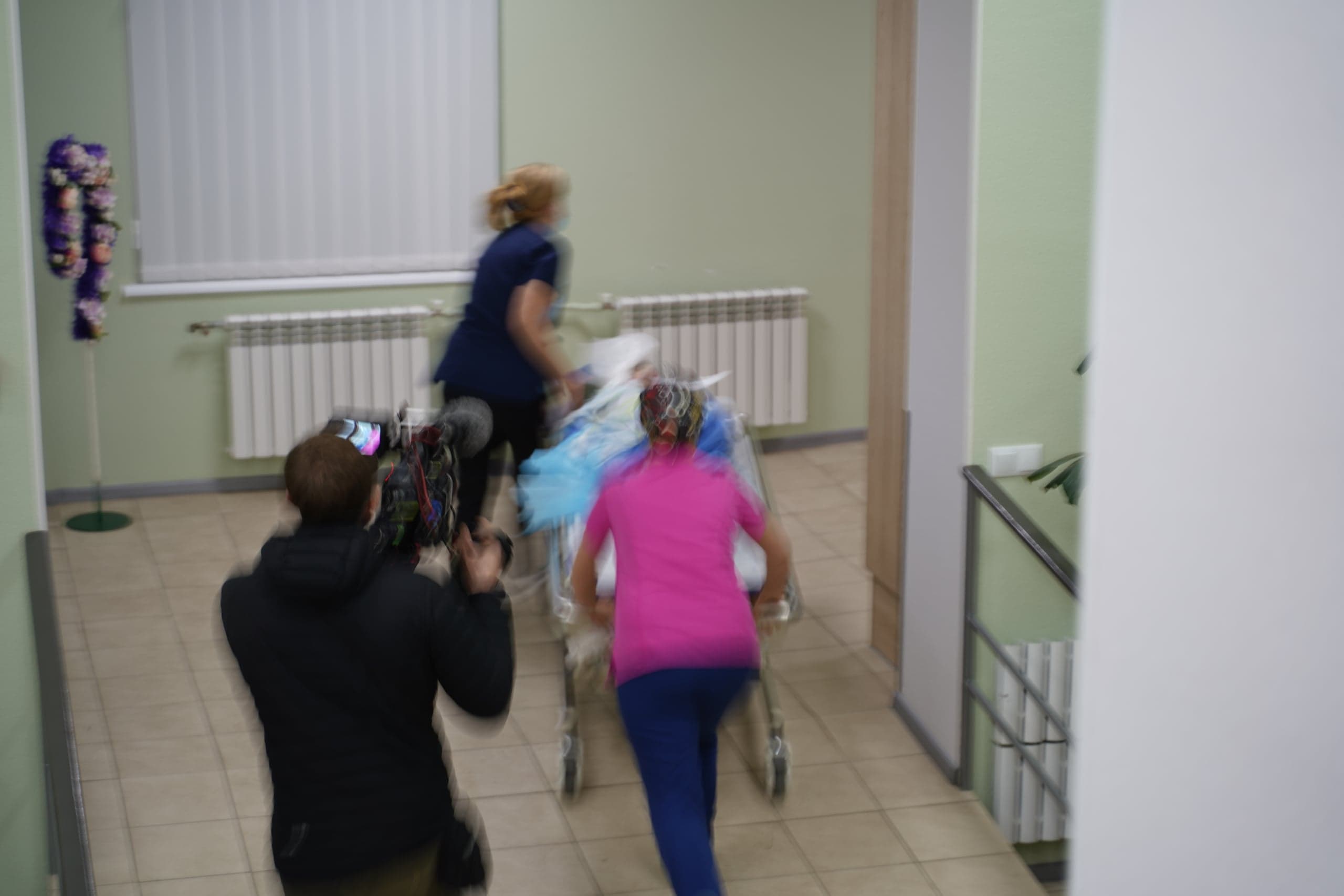 Размытый кадр: оператор снимает двух медработников, быстро везущих пациента на каталке в больничном коридоре.