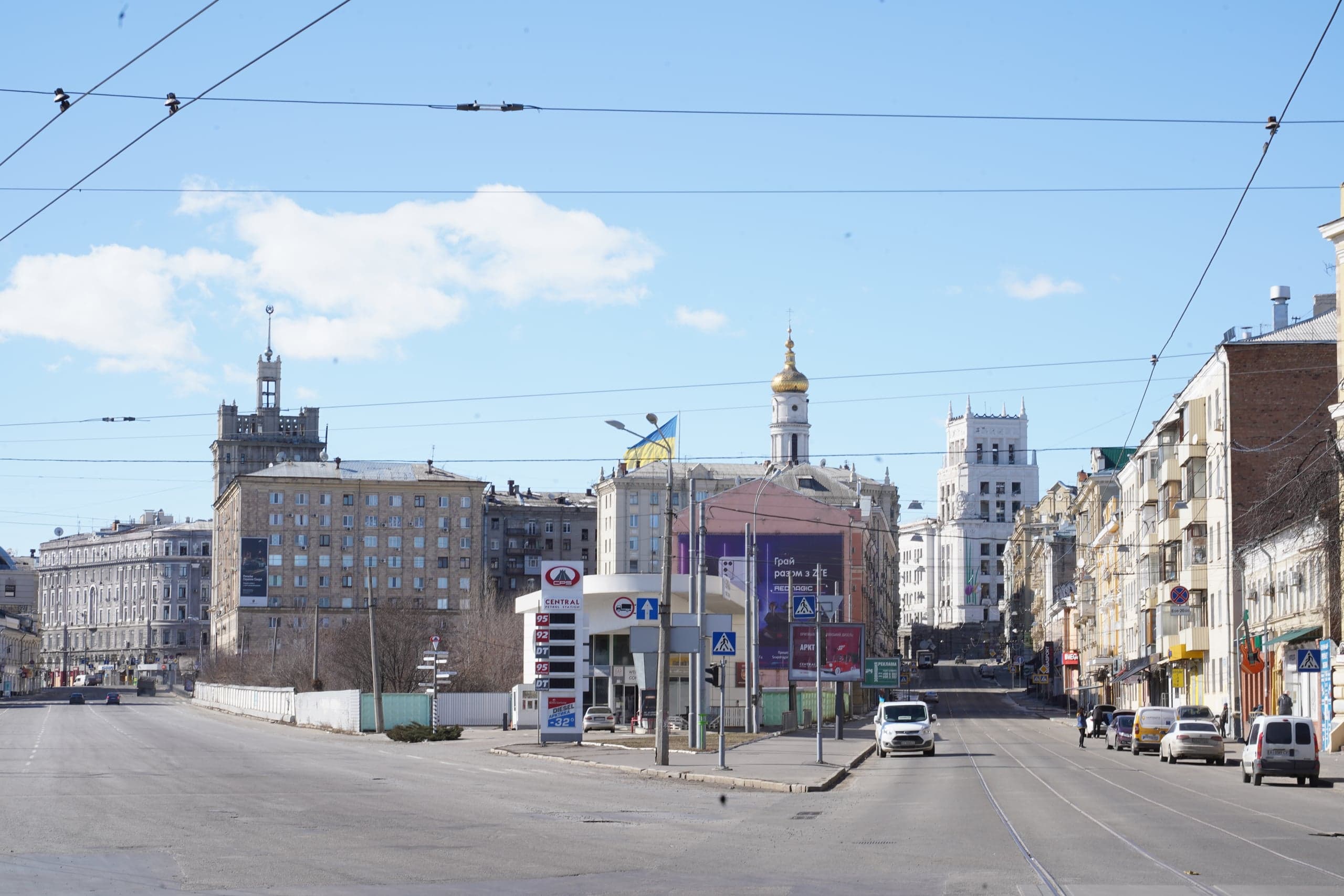 Городской пейзаж: широкий перекресток, окруженный историческими зданиями, включая башню с золотым куполом и сталинский шпиль, под проводами.