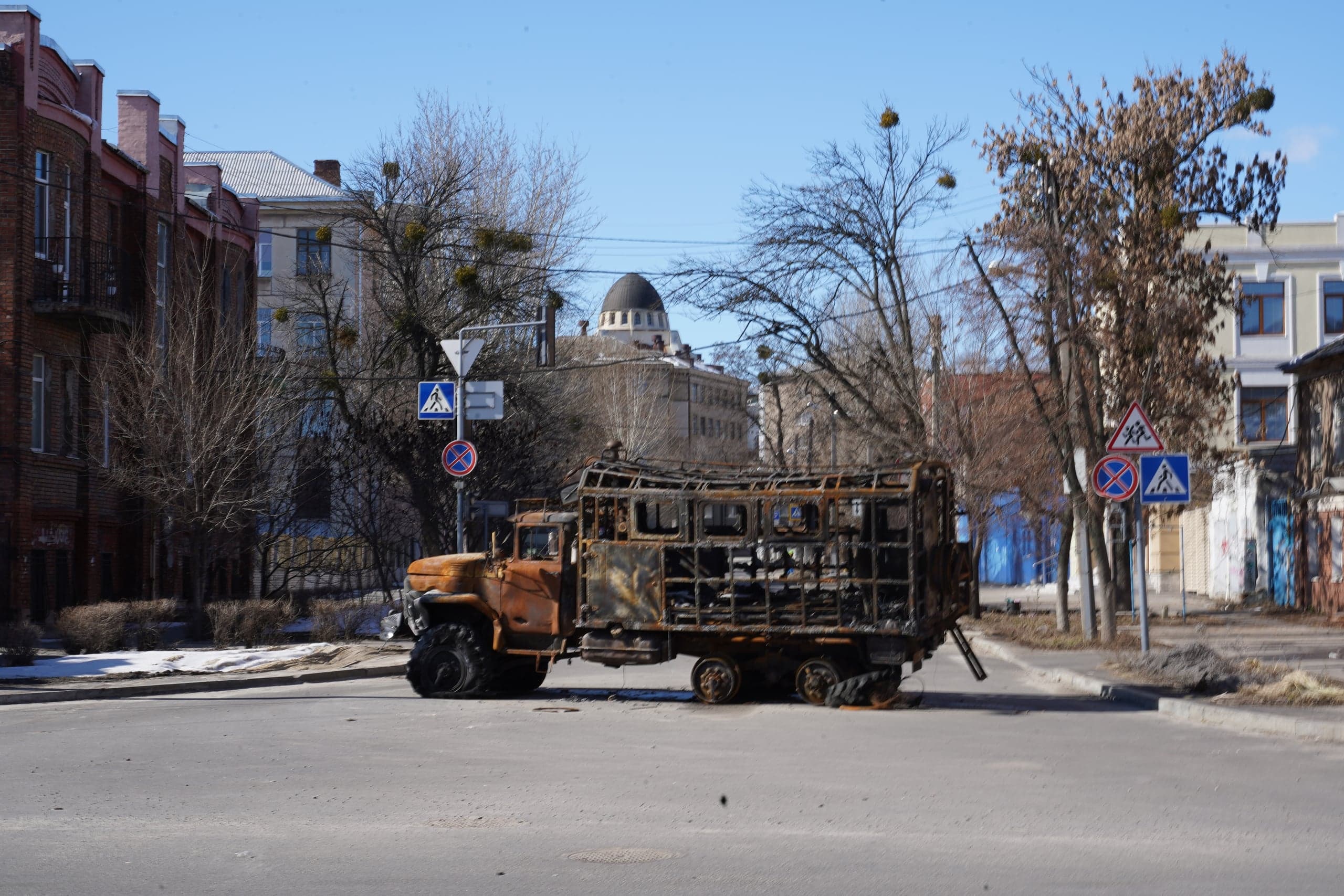 Сильно обгоревший и ржавый остов грузовика (будки) с пробитыми шинами стоит посреди городского перекрестка. Вокруг видны старые здания и голые деревья под голубым небом.