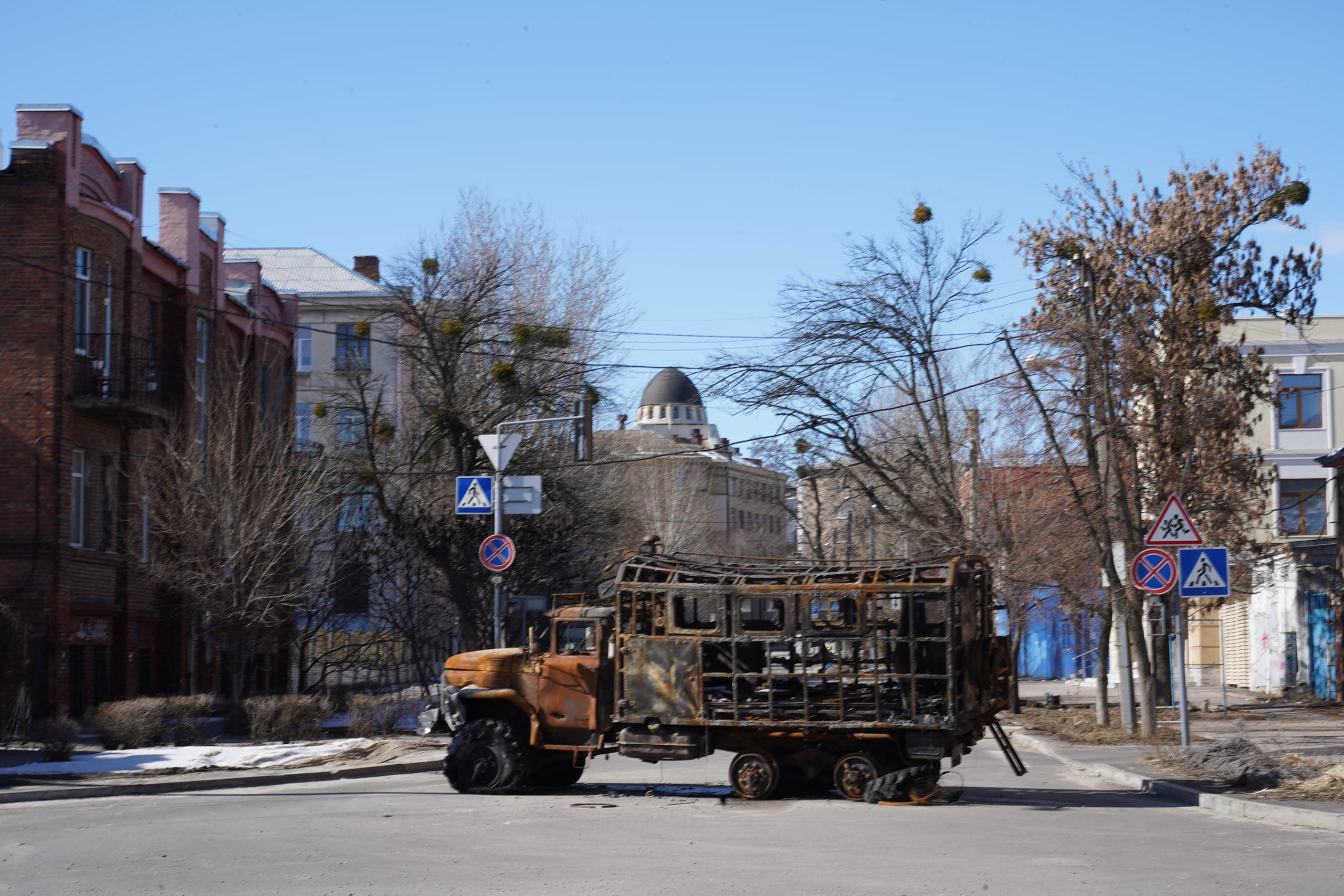 Сгоревший, покрытый ржавчиной грузовик с уничтоженными колесами стоит на перекрестке в городском квартале с историческими зданиями и голыми деревьями. Ясное небо.