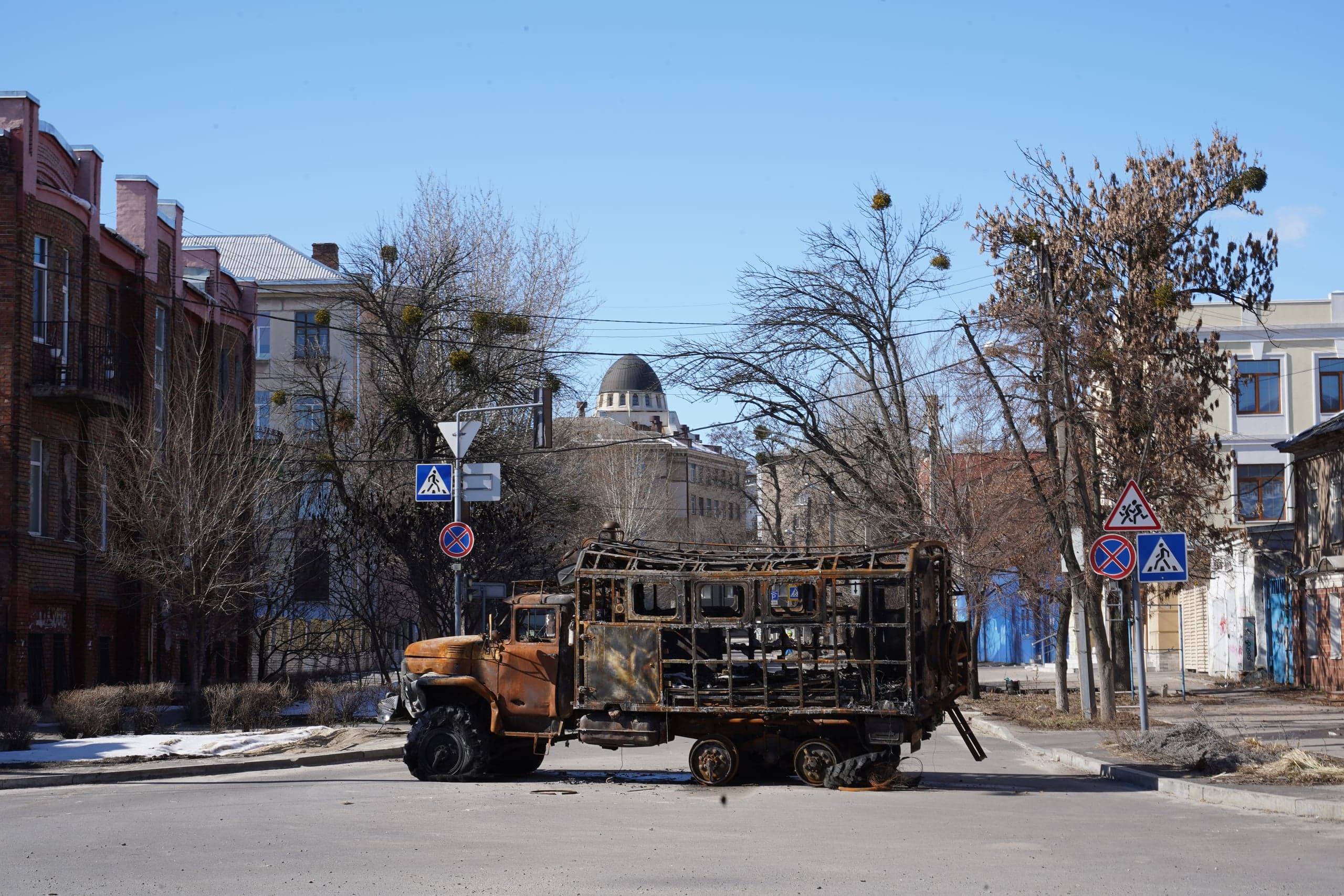 Остов сгоревшего и ржавого грузовика стоит на городской улице между жилыми домами и голыми деревьями под голубым небом.