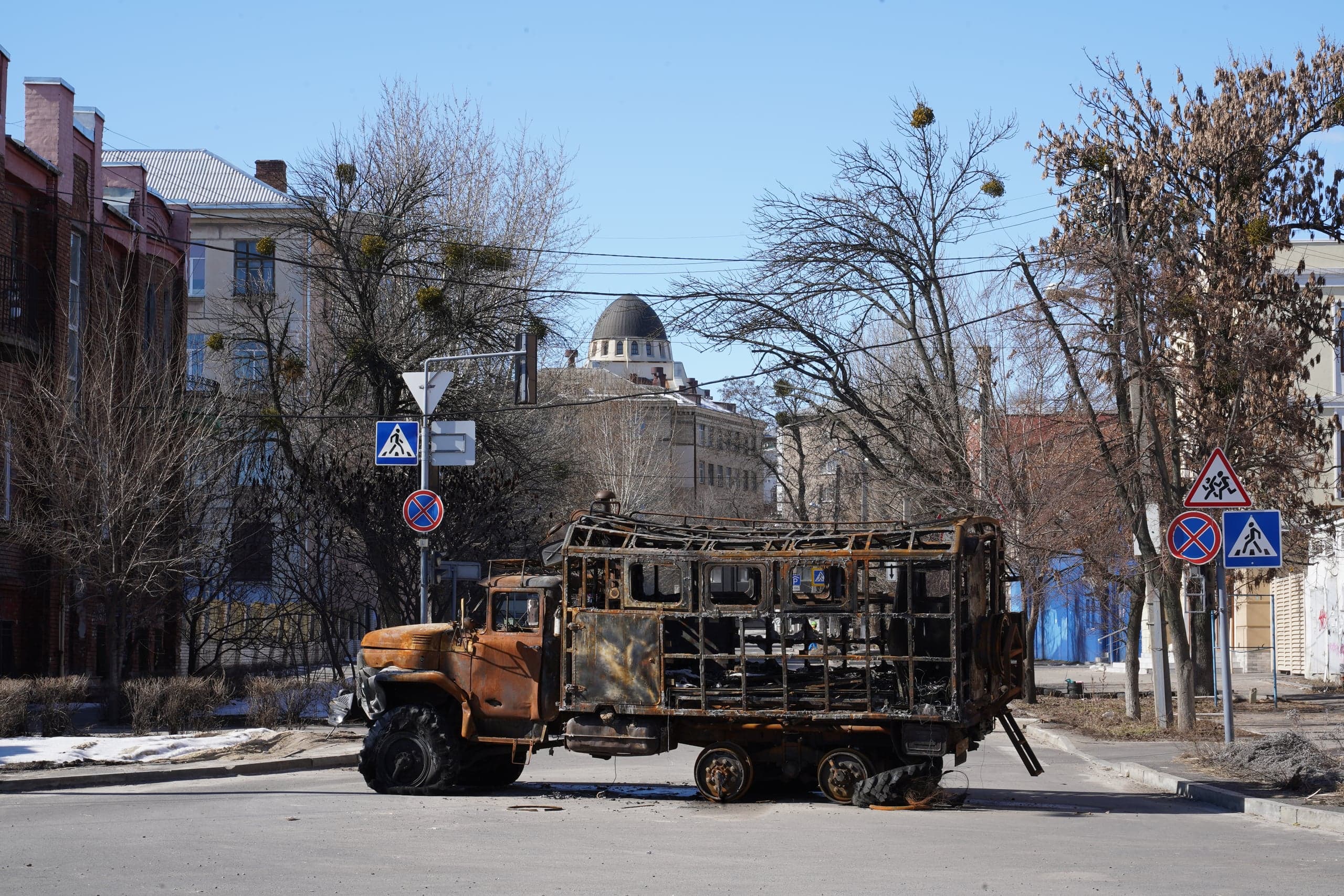 Сгоревший и ржавый остов грузовика стоит посреди асфальтированной городской улицы. На заднем плане видны жилые здания и купол собора под голубым небом.