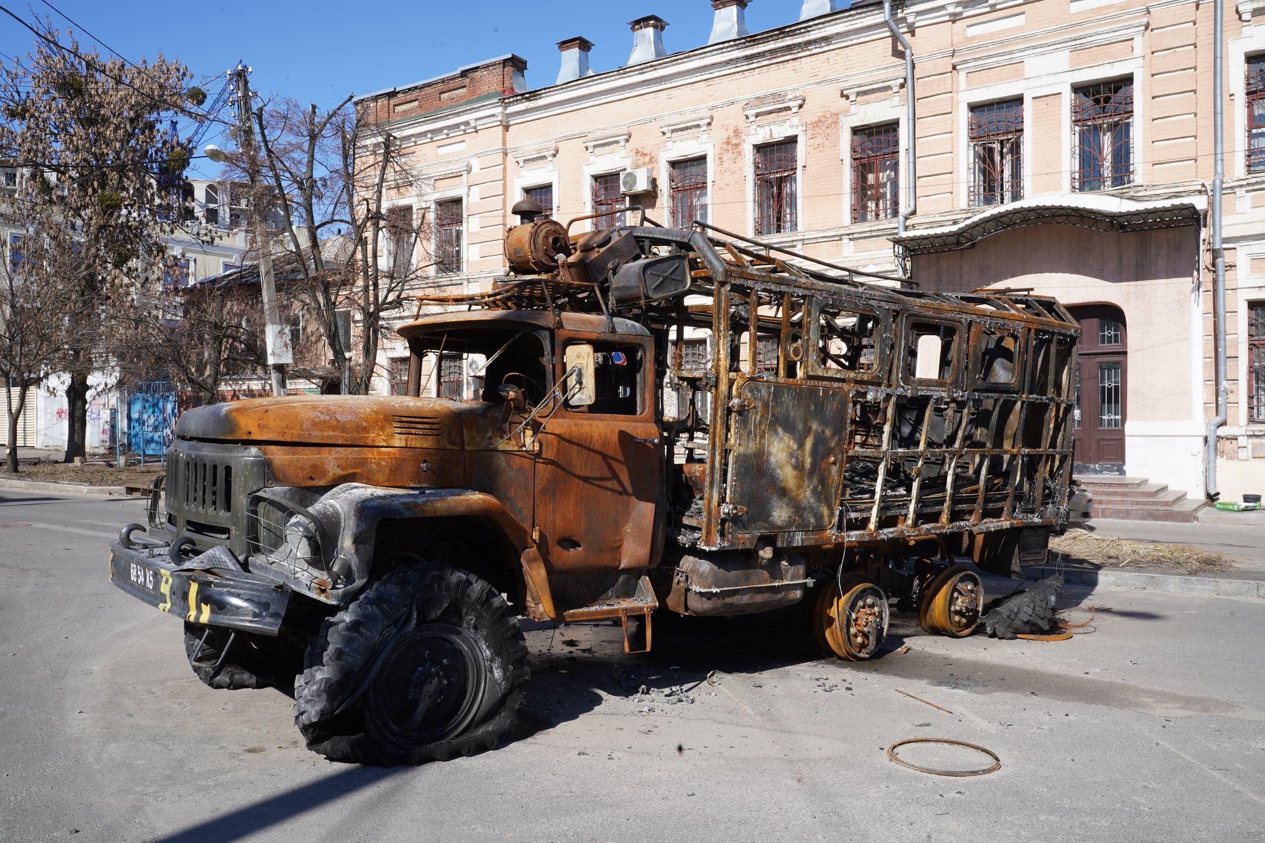Сгоревший, ржавый остов военного грузового автомобиля (кузов превращен в каркас) стоит на городской улице на фоне старинного светлого здания.