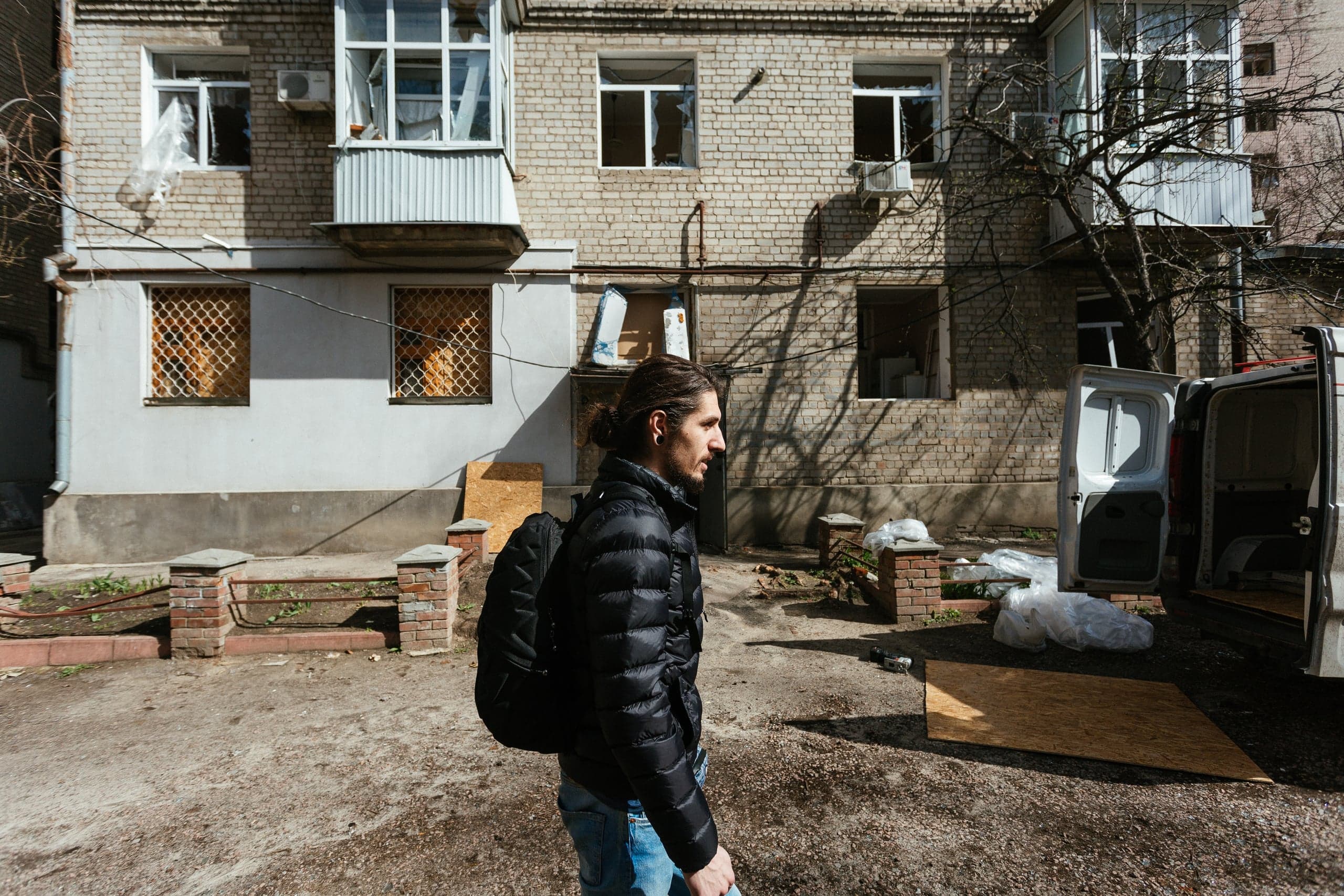 Мужчина с рюкзаком идет мимо поврежденного кирпичного дома с разбитыми окнами и открытым фургоном.