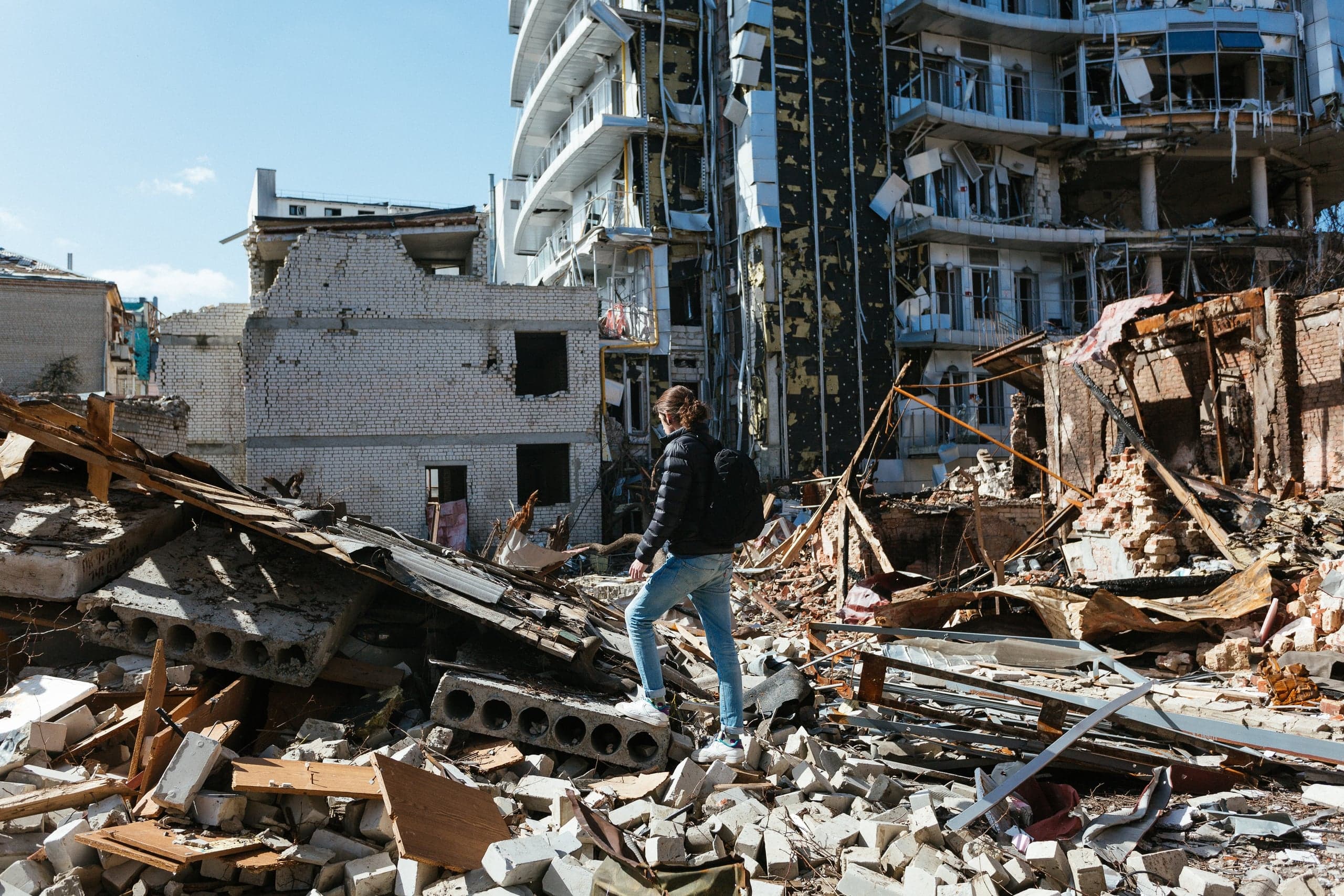 Человек стоит на груде строительных обломков на фоне сильно разрушенных многоэтажных зданий.