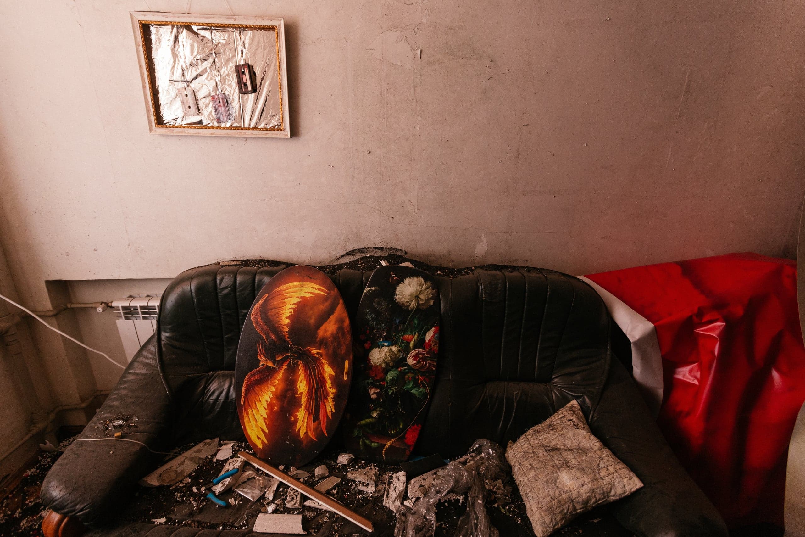 Черный разрушенный диван в грязной комнате, заваленный обломками. На спинке стоят две декоративные доски (с огненными крыльями и цветами). На стене слева — рамка с фольгой и кассетами.