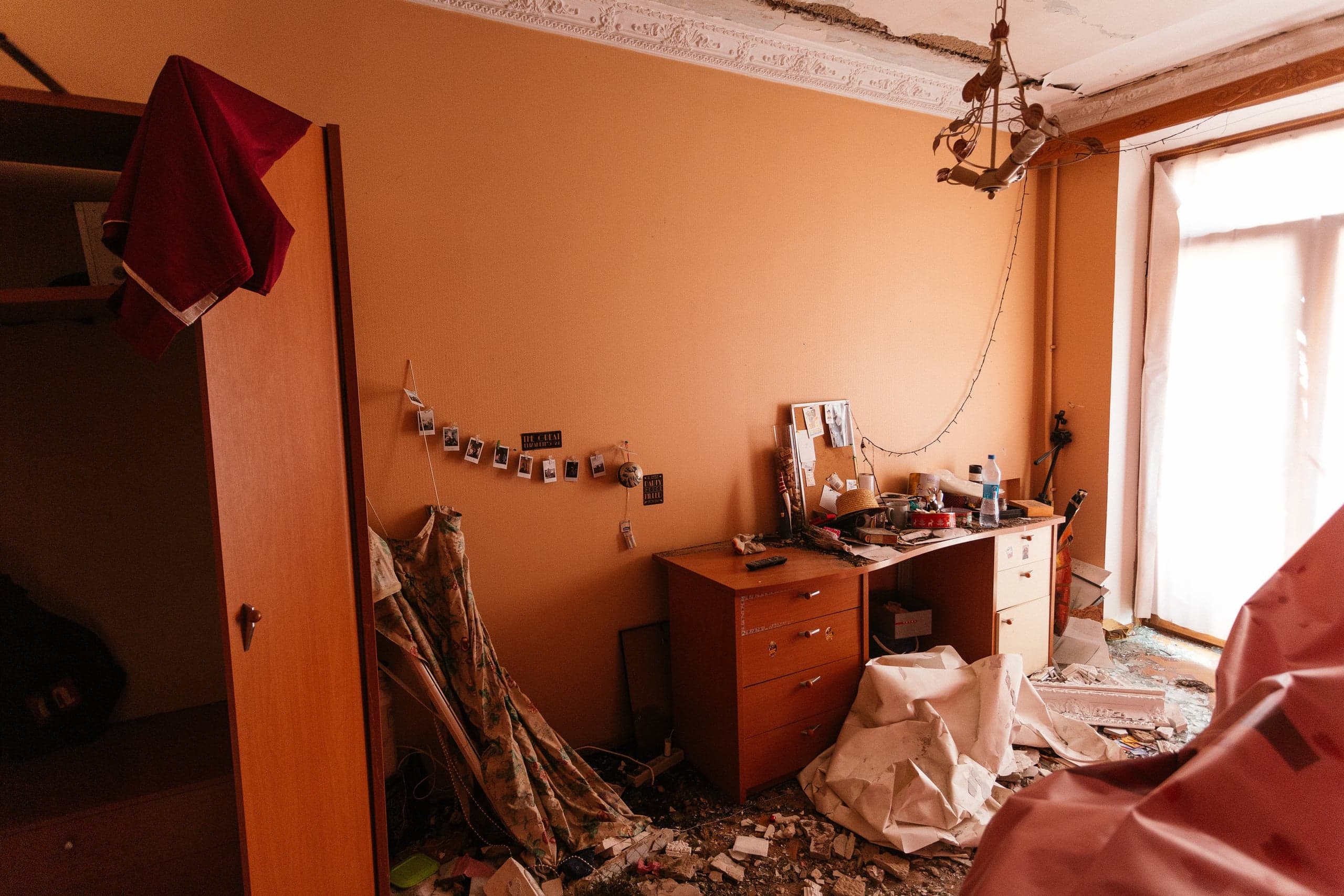 Разрушенная комната: персиковые стены, обвалившаяся потолочная лепнина и висящая люстра. Пол покрыт обломками и строительным мусором. У стены стоит захламленный стол с личными фотографиями; справа яркий свет льется из пересвеченного оконного проема.