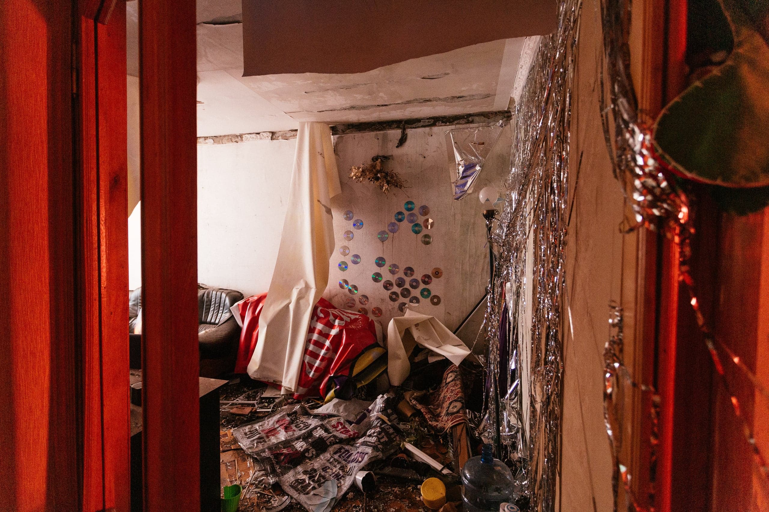 Вид через красные дверные косяки на захламленную, полуразрушенную комнату. На потрескавшейся стене приклеены CD-диски, а правая стена покрыта серебряным дождиком. На полу строительный мусор и наваленная ткань.