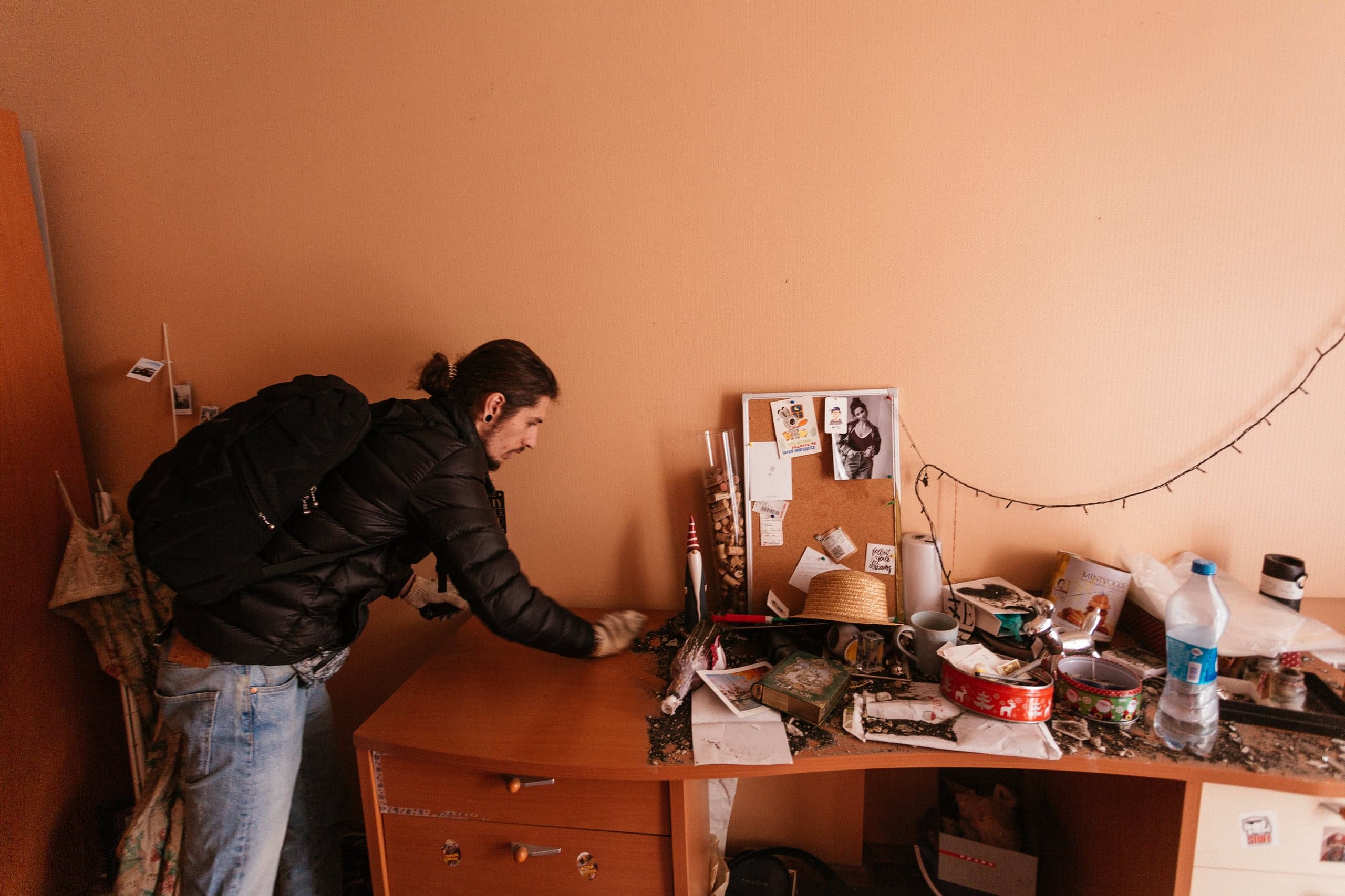 Мужчина в черной куртке и рюкзаке вытирает темный мусор с заваленного личными вещами рабочего стола.