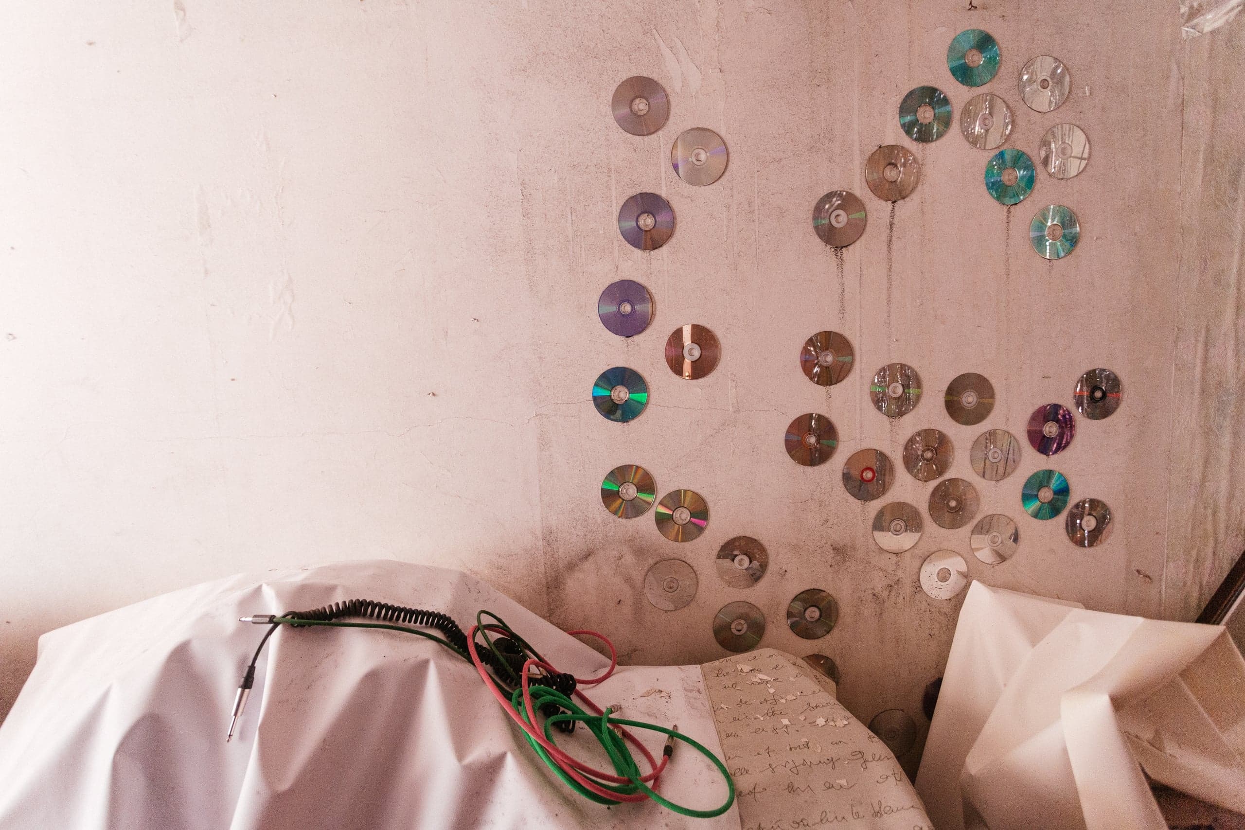 Стенд из CD-дисков на грязной стене, внизу — аудиопровoда на белой ткани и старые рукописи.